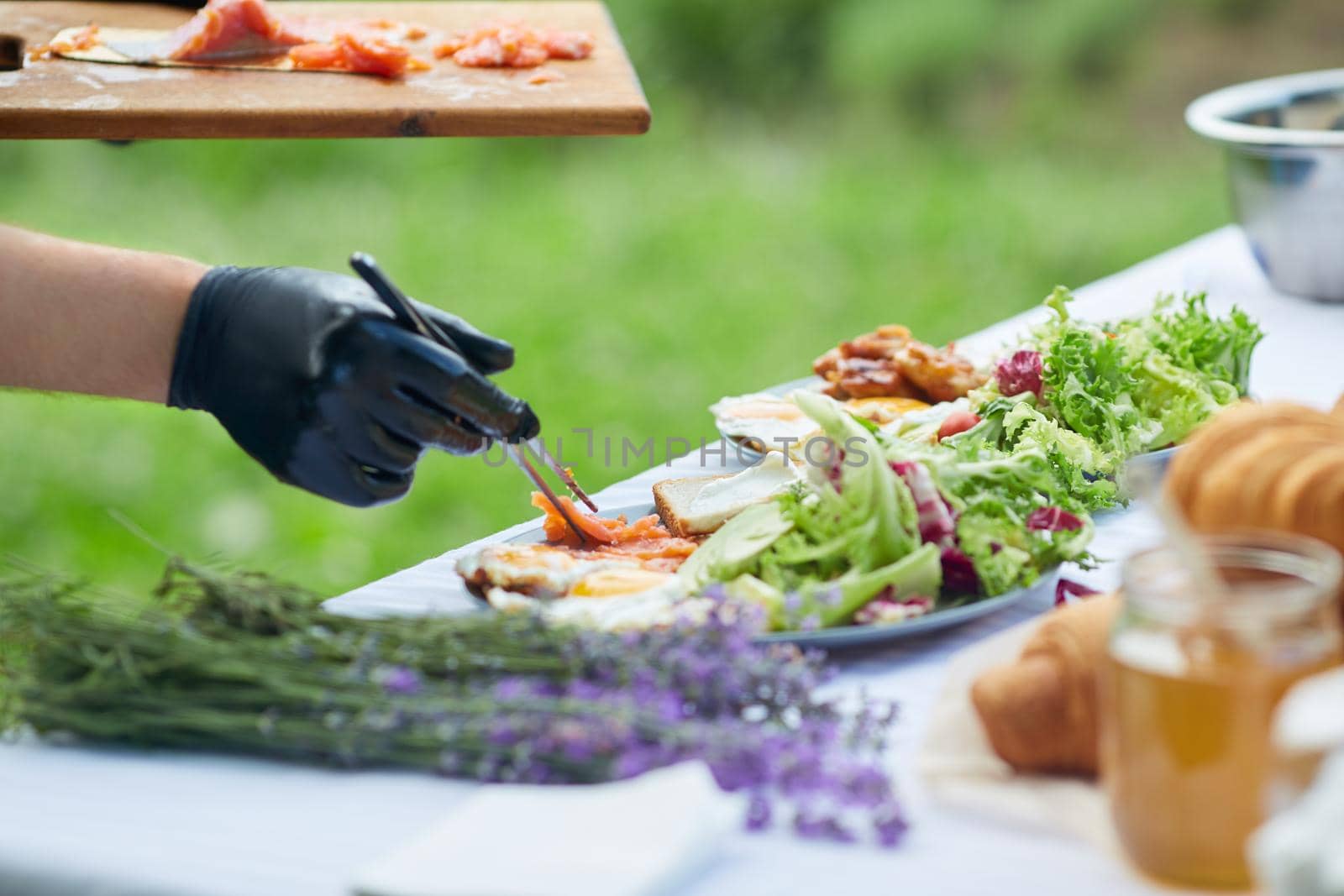 Male hands in gloves putting salmon from cutting board using iron twizzers. Side view of unrecognizable chef finishing dish, bread and fresh salad on plates, table decorated with lavender bouquet.