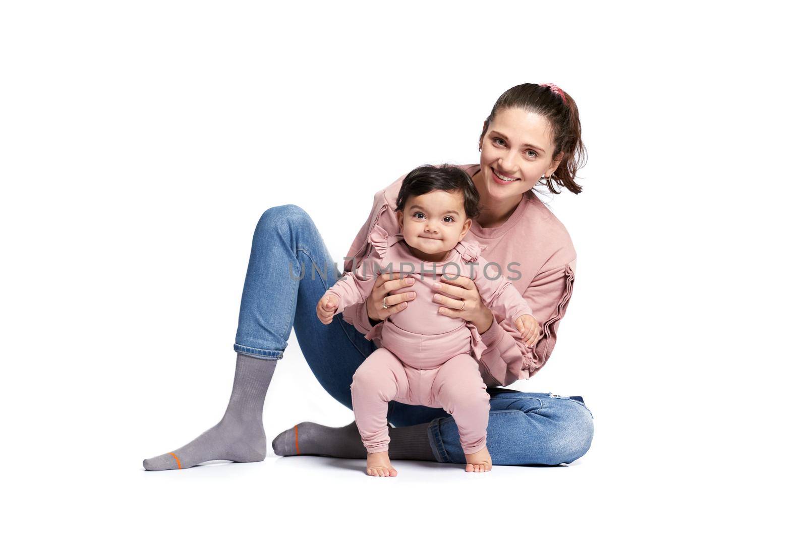 Portrait of cute mother with daughter looking at camera isolated on white studio background. Young attractive woman sitting on floor and helping sweet adorable child to stand, happy childhood concept.