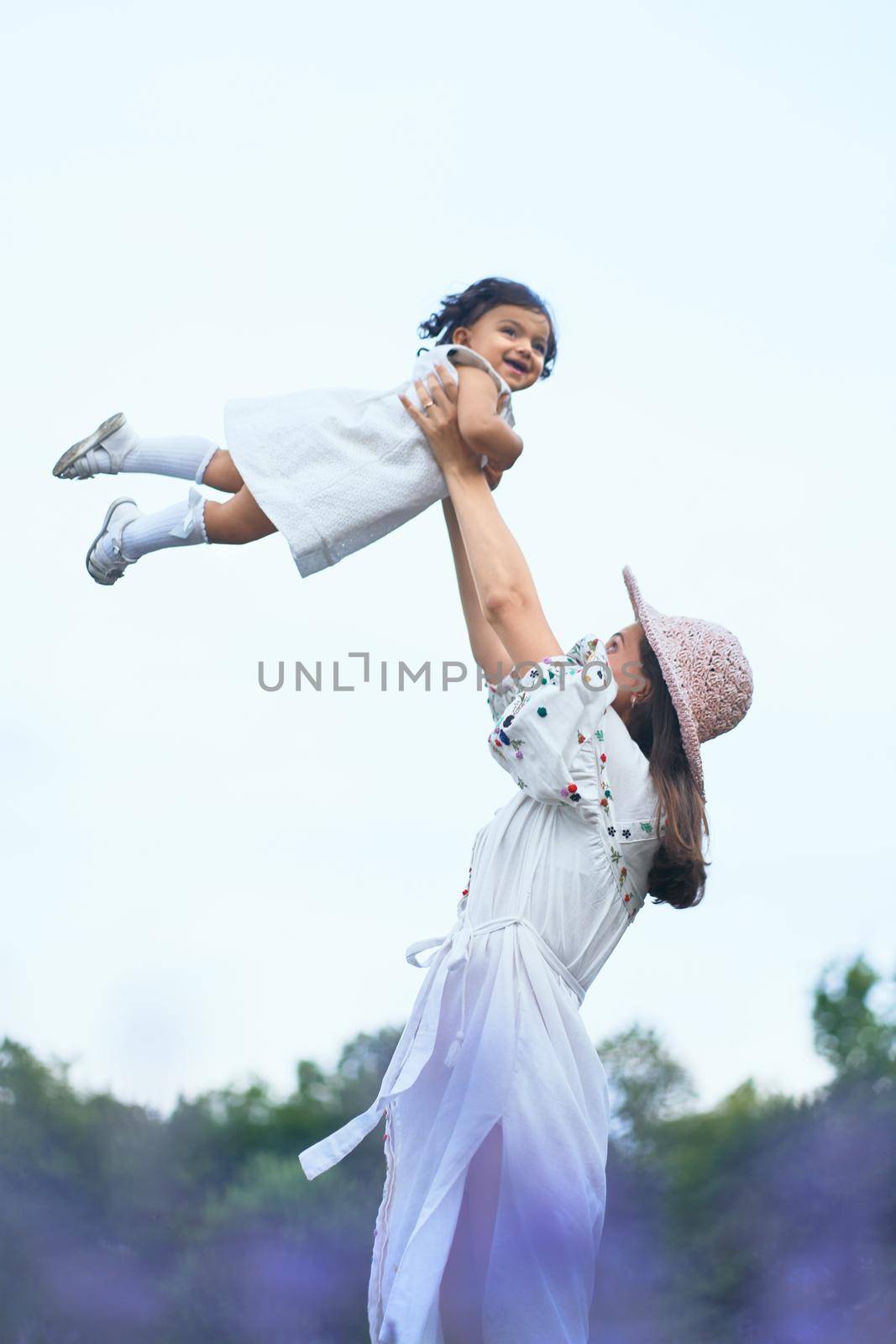 Young loving mother wearing straw hat enjoying time with little baby daughter, playing with laughing kid. Stunning lady wearing dress throwing child up, blooming lavender field on background.