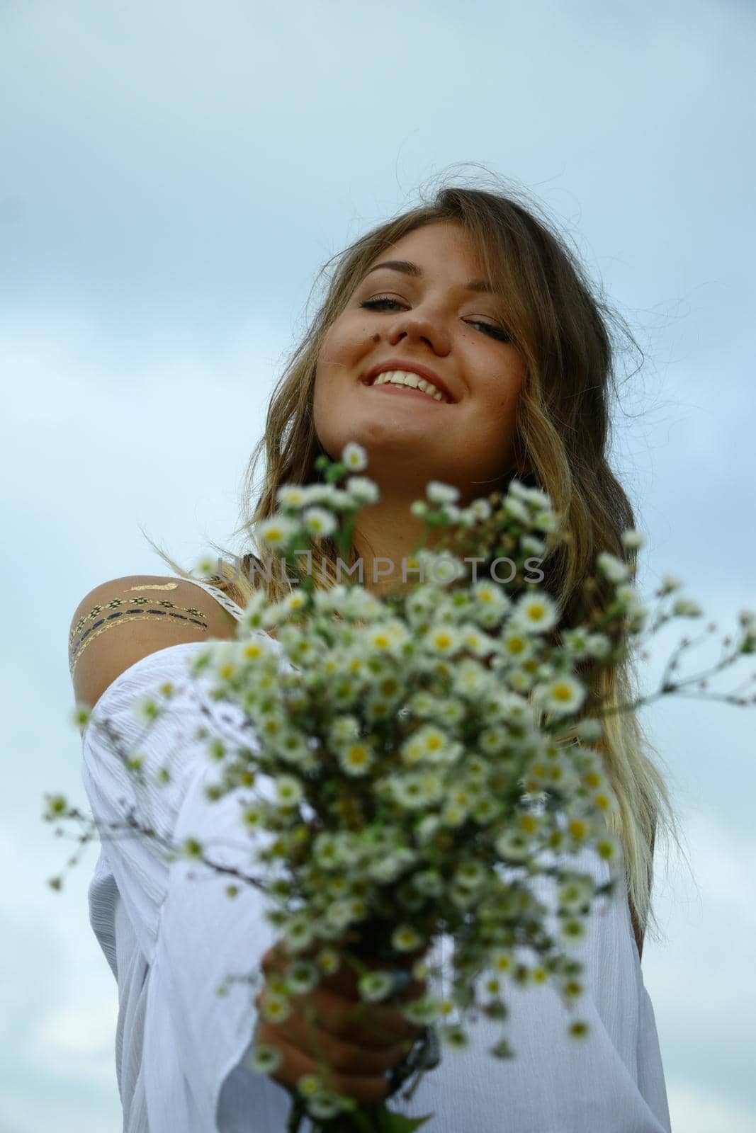 portrait of a happy flirty beautiful blonde woman in white blouse in the field. Carrying a bouquet of daisies. Temporary tattoo. Drawings on body. hippie. Nature loving.