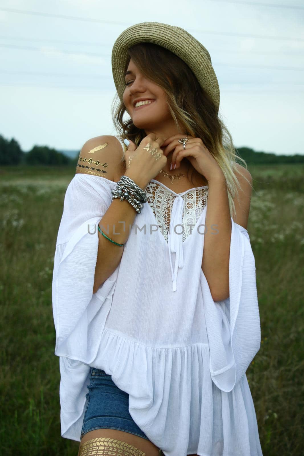 portrait of a happy flirty beautiful blonde woman in white blouse and straw hat in the field. Temporary tattoo. Drawings on body. hippie. Nature loving.