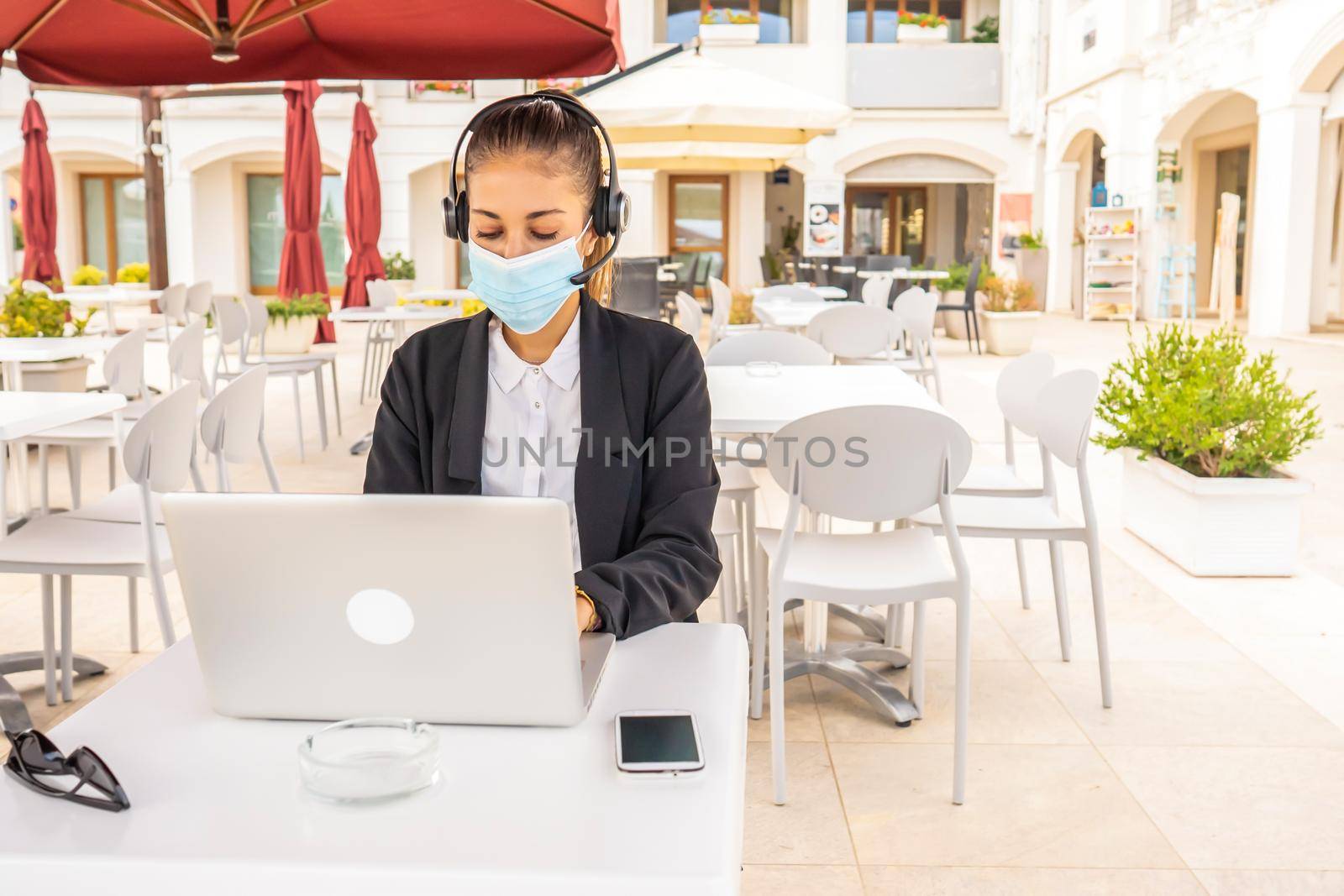 Self entrepreneur woman working in outdoor bar table using laptop wearing protective medical face mask due to social distancing for Covid-19 pandemic. New normal jobs with mobile wi-fi technology by robbyfontanesi