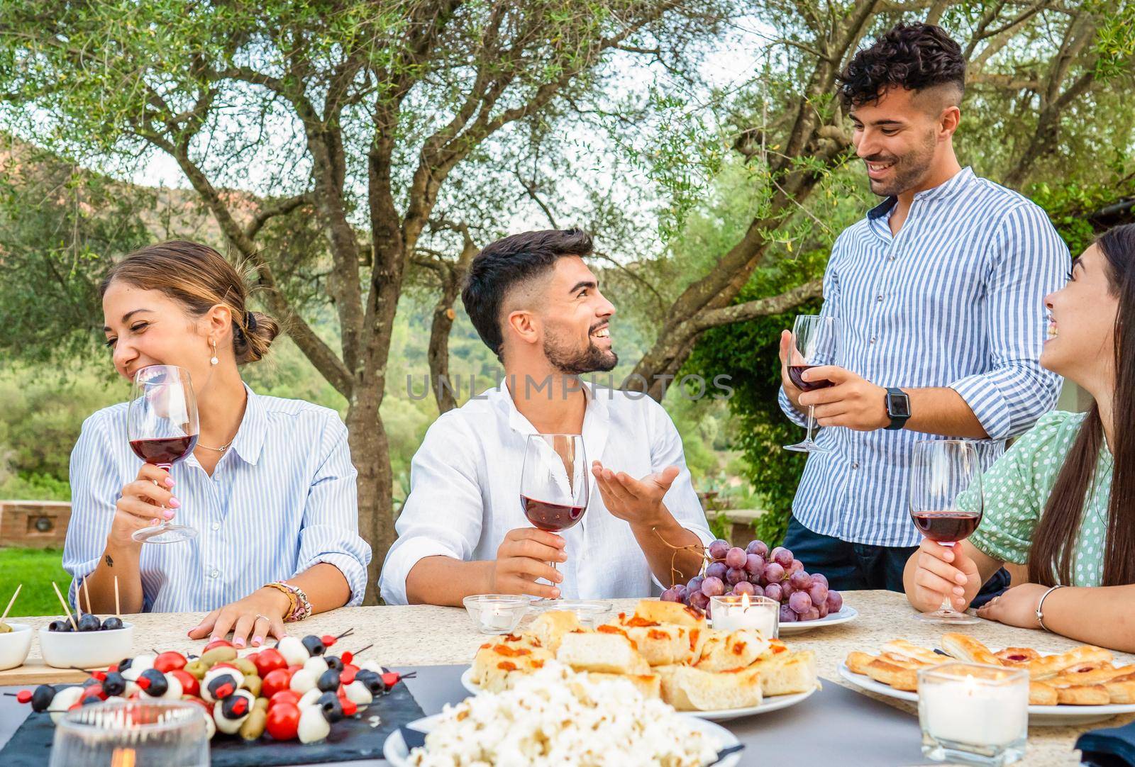 Typical italian celebration for grape harvest in country size house. Group of young friends gathering at table laden with snacks, pizza, tomatoes and mozzarella skewers holding glasses of red wine