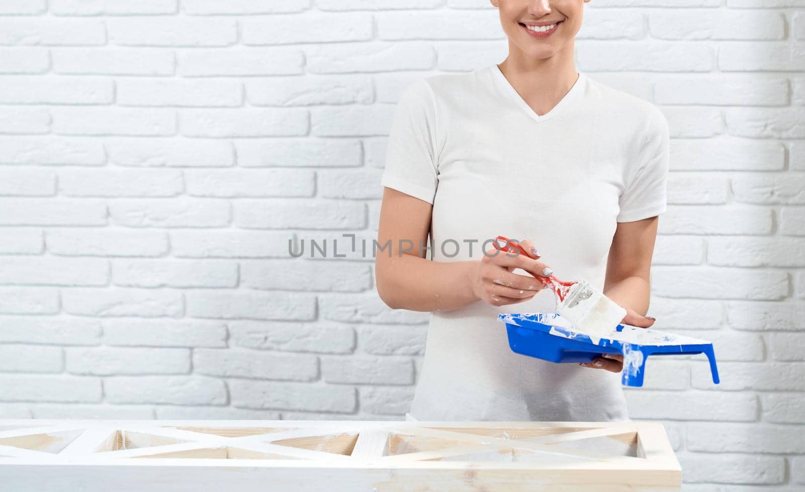 Smiling woman in white shirt holding paint on white background. Concept of preparing for painting wooden rack.