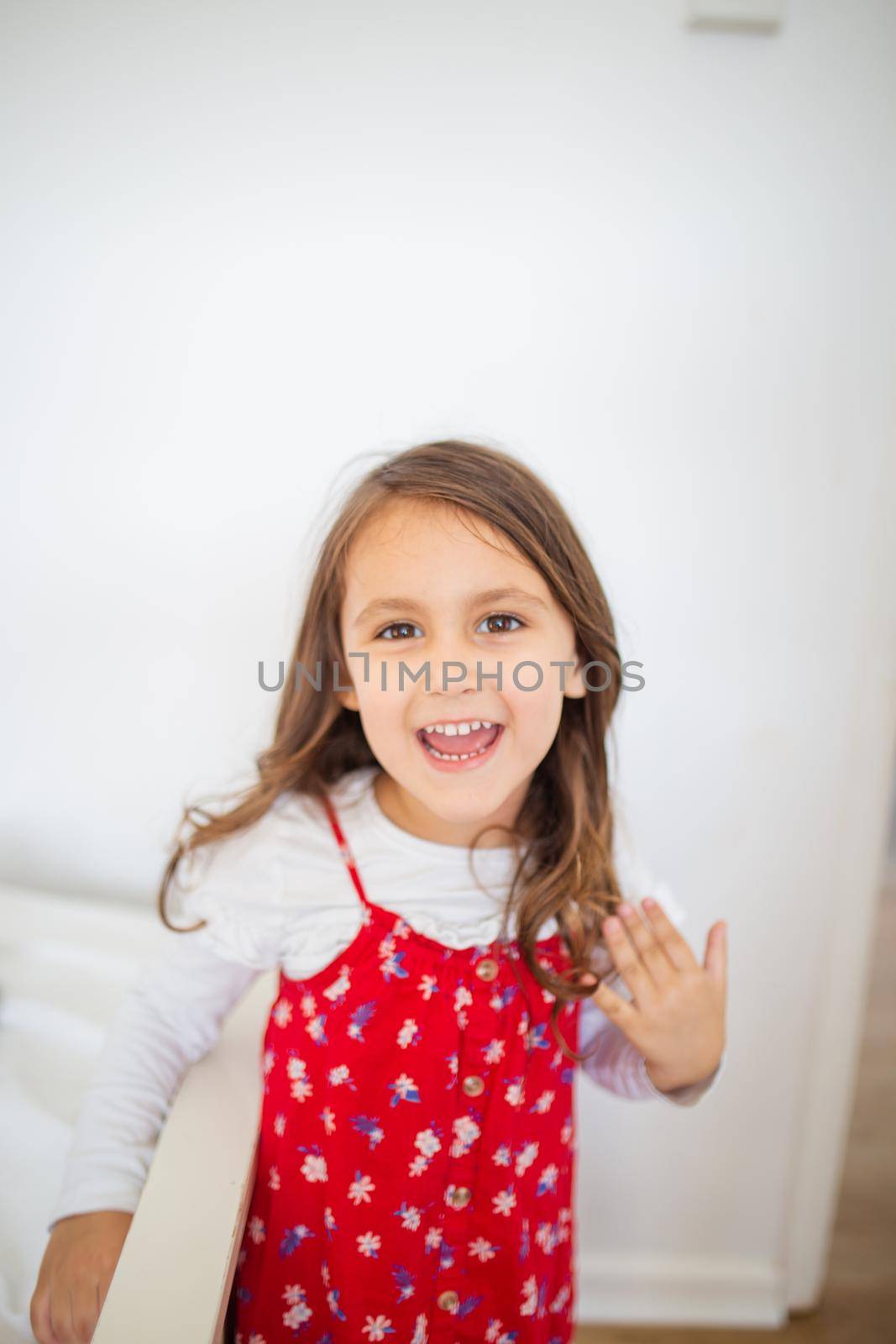 Cute little girl inside white room smiling and wearing red-flowered dress. Portrait of joyful young child in front of white wall. Happy kids at home