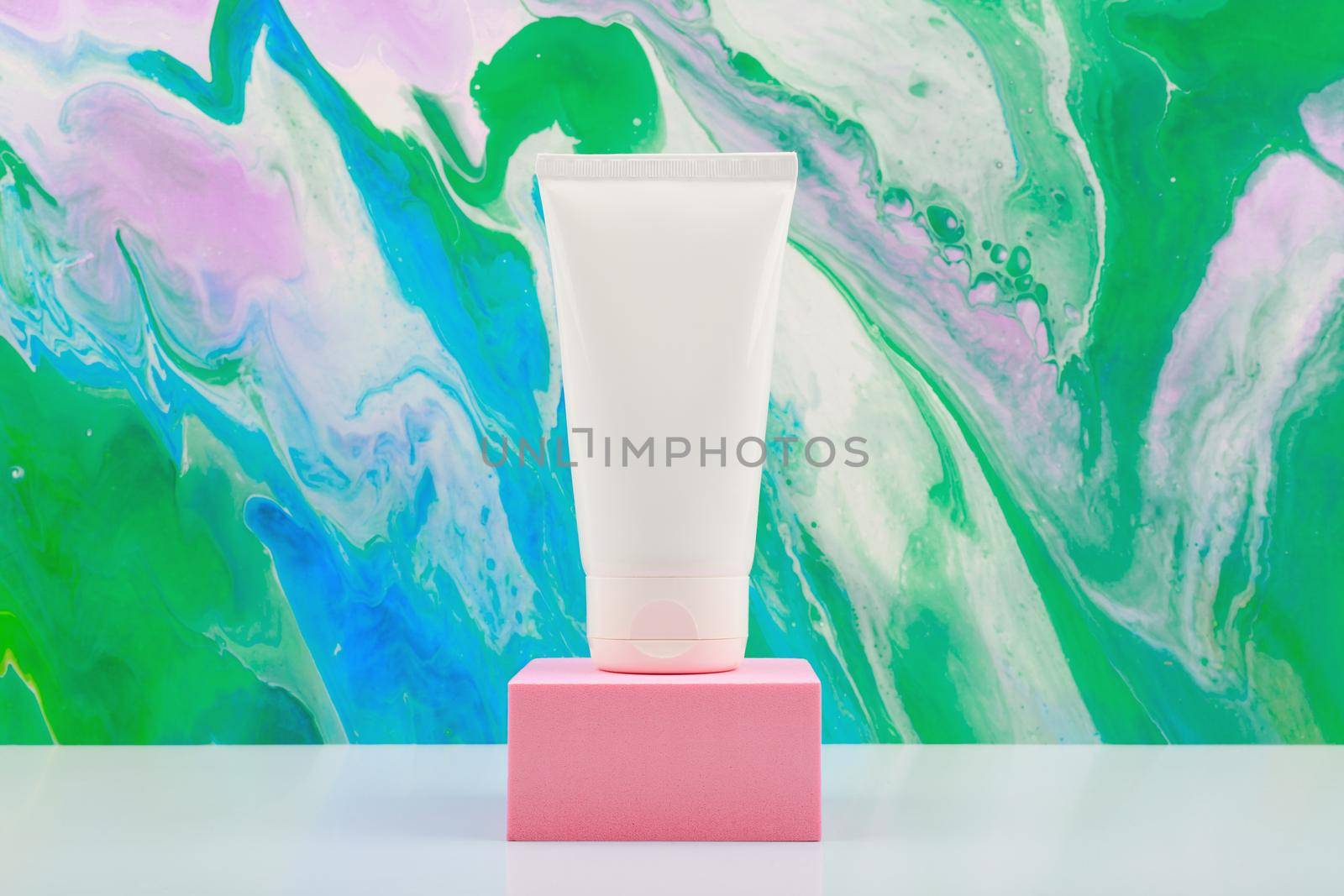 Hands cream in white unbranded tube on pink podium against bright marbled background in green and blue tones. Concept of groomed hands and soft, moisturized skin