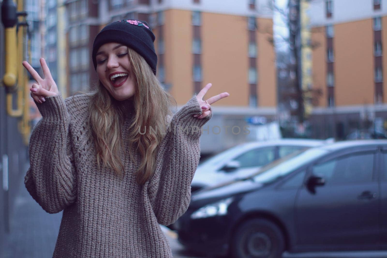 urban portrait of a happy flirty beautiful woman in knitted brown sweater, jeans and black hat. in the parking lot.