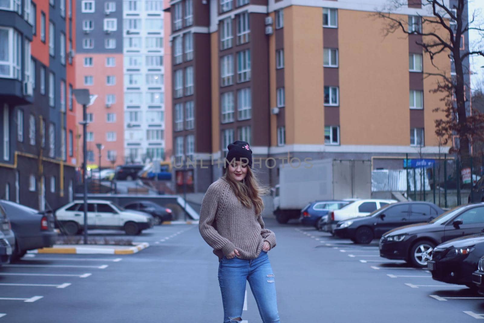 urban portrait of a happy flirty beautiful woman in knitted brown sweater, jeans and black hat. in the parking lot.