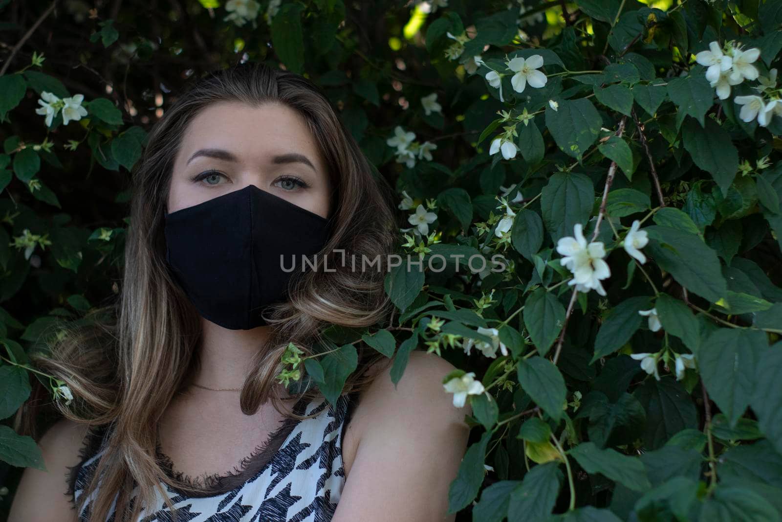 Pretty young blonde woman in medical black face mask. in a bush with little white flowers. modern reality. covid-19 concept. copy space. High quality photo