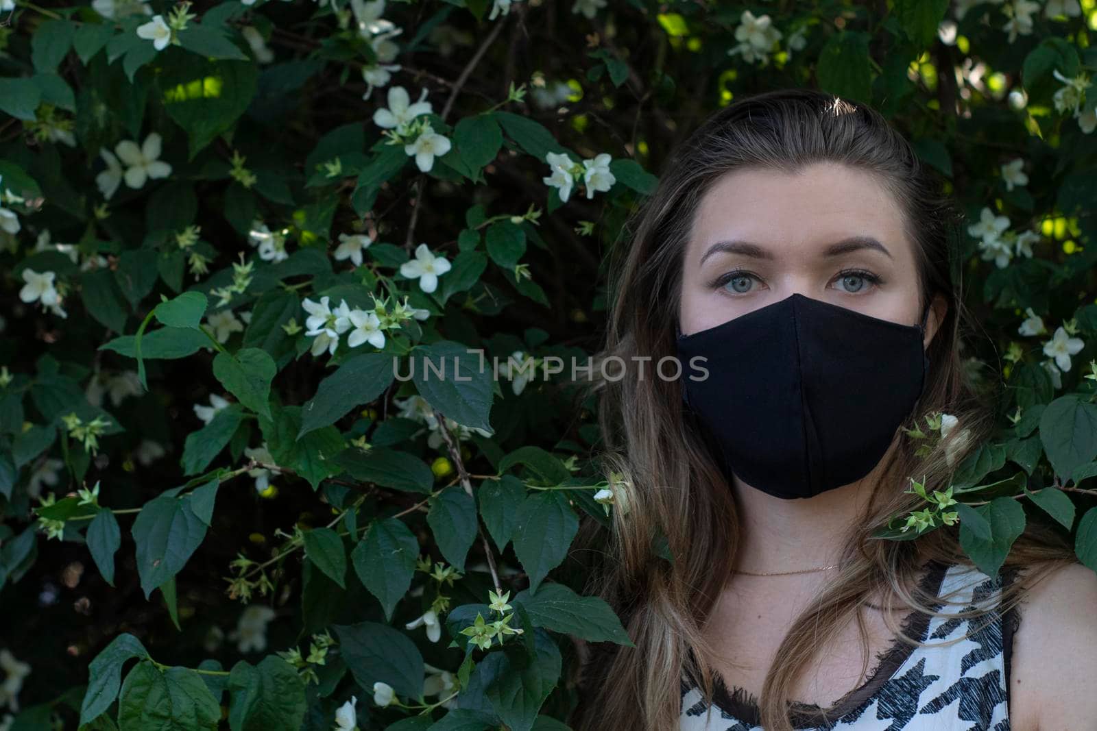 Pretty young blonde woman in medical black face mask. in a bush with little white flowers. modern reality. covid-19 concept. copy space. High quality photo