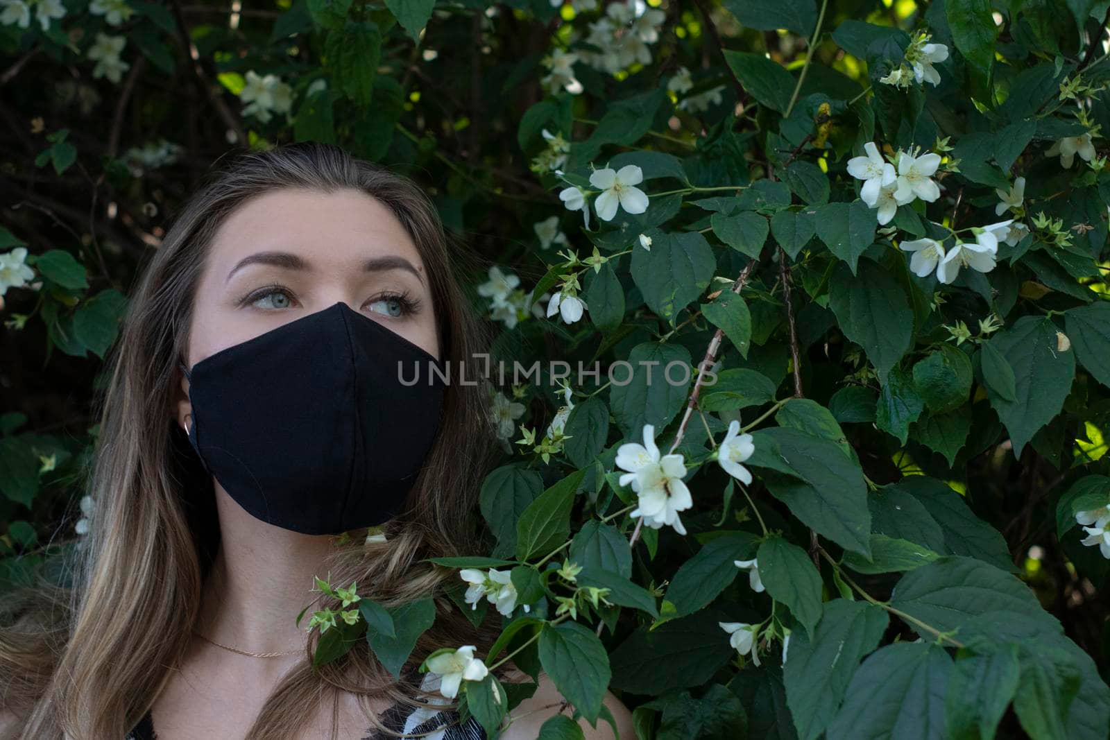 Pretty young blonde woman in medical black face mask. in a bush with little white flowers. modern reality. covid-19 concept. copy space. High quality photo