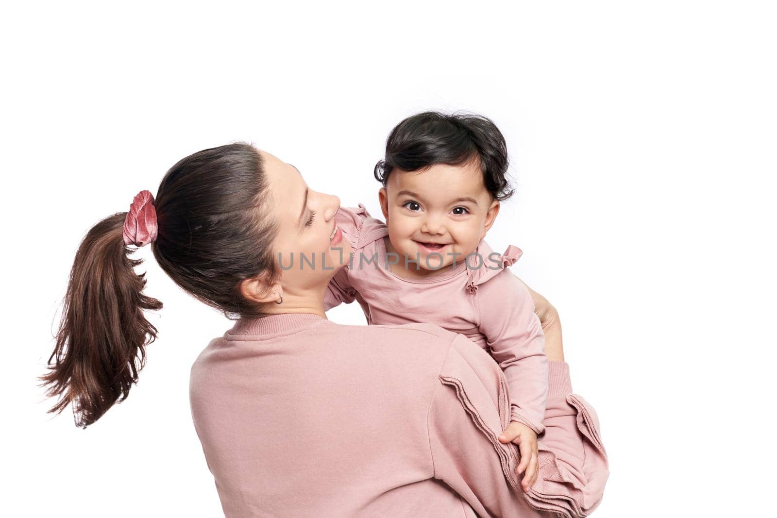 Back view of young mother having fun with daughter, isolated on white studio background. Young woman hugging sweet adorable child, baby girl looking at camera, happy childhood concept.