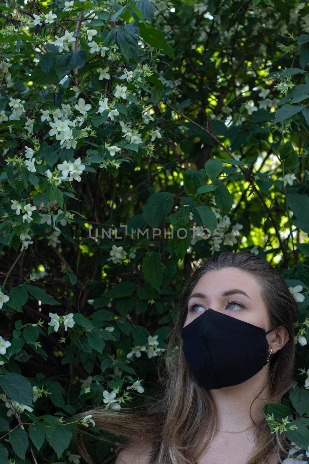 Pretty young blonde woman in medical black face mask. in a bush with little white flowers. modern reality. covid-19 concept. copy space. High quality photo