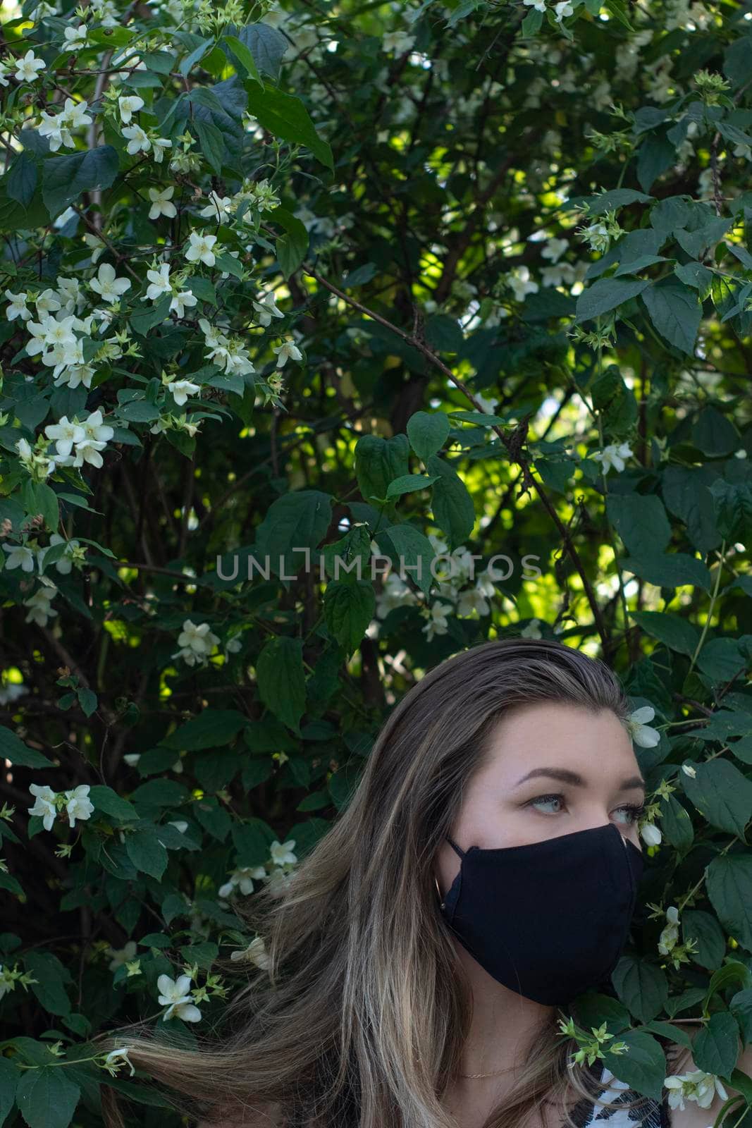 Pretty young blonde woman in medical black face mask. in a bush with little white flowers. modern reality. covid-19 concept. copy space. High quality photo