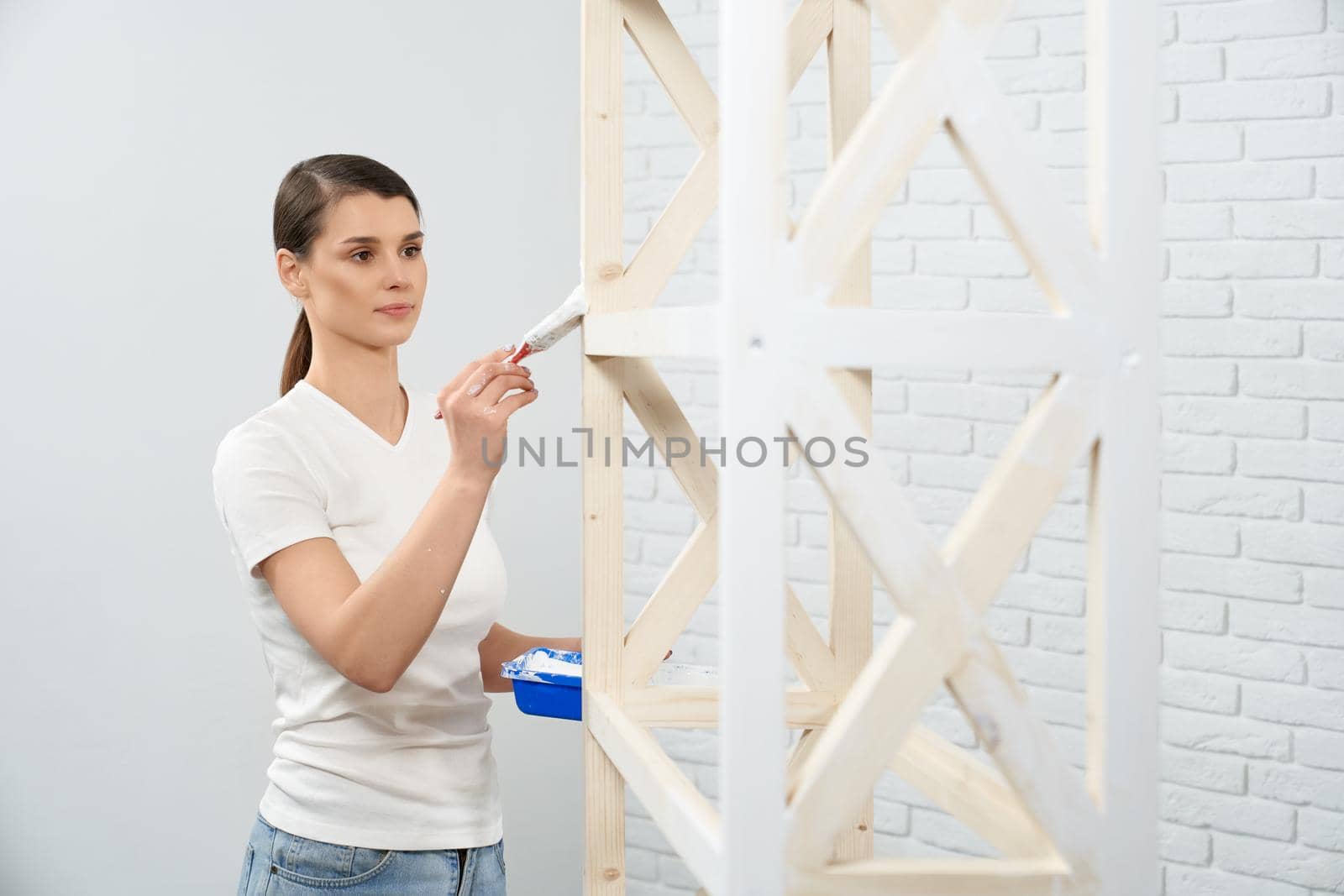 Brunette woman painting wooden rack in empty room. by SerhiiBobyk