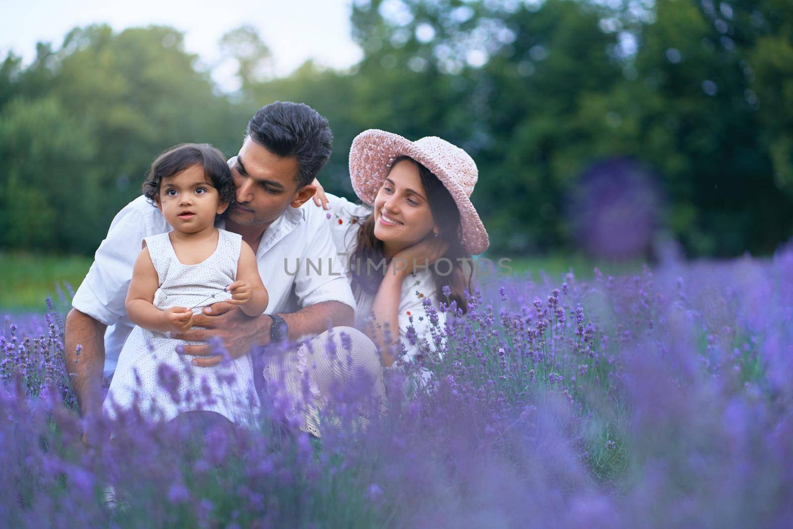 Front view of mother, father and daughter sitting in lavender field and smiling. Cute baby girl, carrying flower and enjoying time with loving parents outdoors. Young family, nature concept.