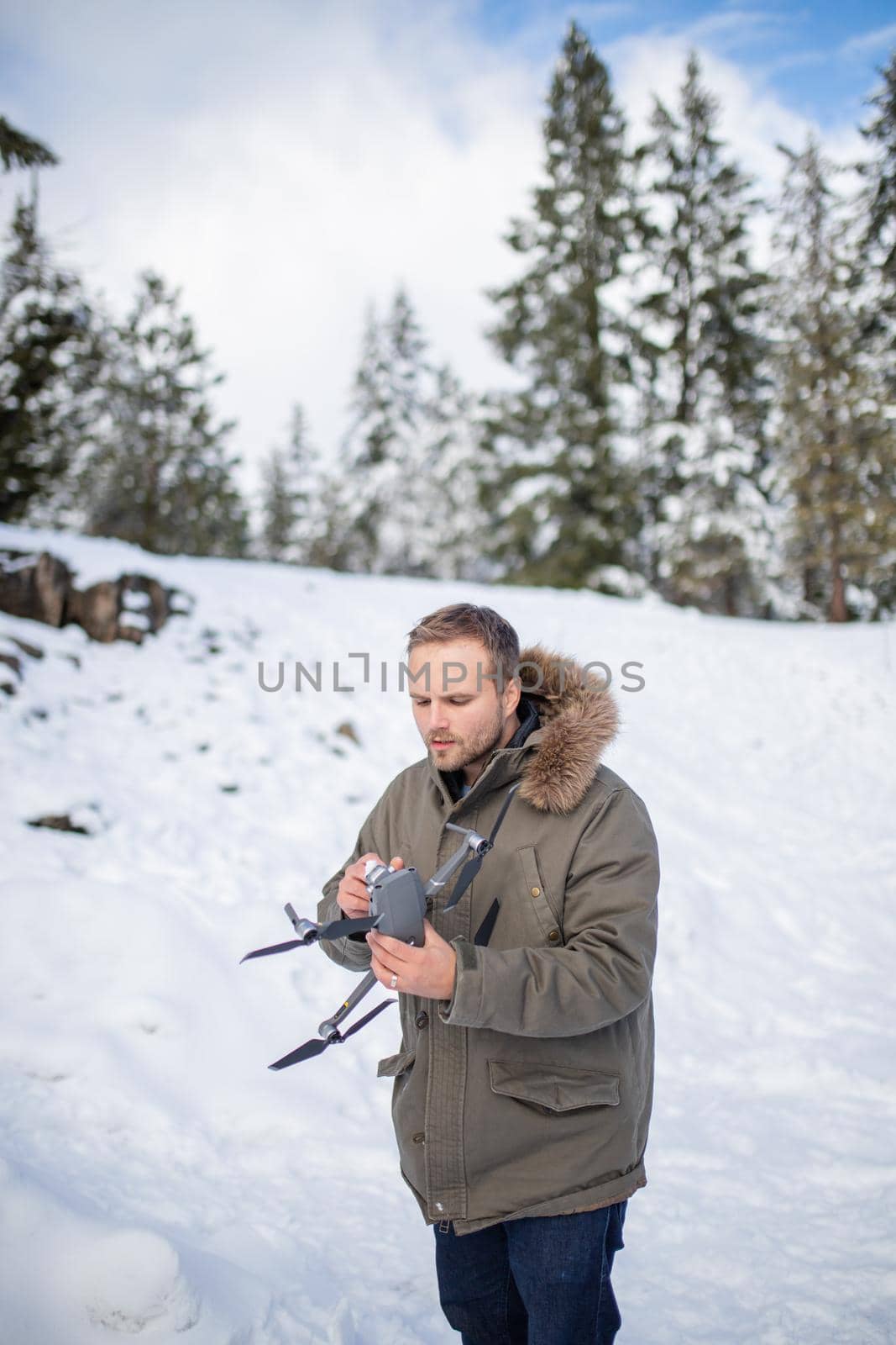 Handsome man holding drone with snowy pines as background. Portrait of concentrated man interacting with drone in frozen woods. Adventurous winter holiday