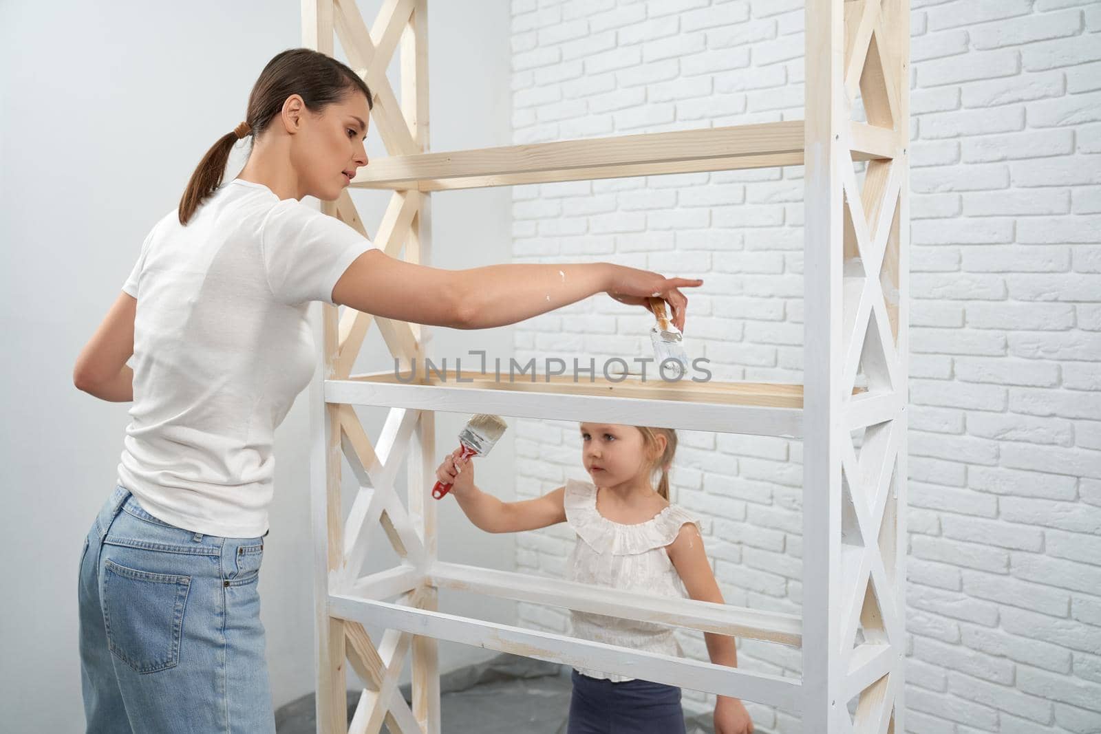 Young mother with daughter together painting rack in white color in bright room. Concept of process painting wooden rack.