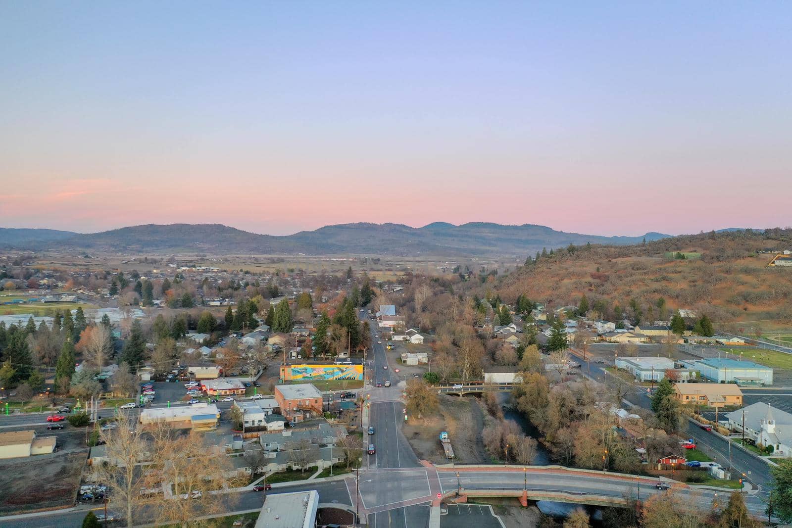 Peaceful town surrounded by trees with mountains as background during sunset. Aerial view of roads surrounded by autumn-colored nature. Rural town landscape