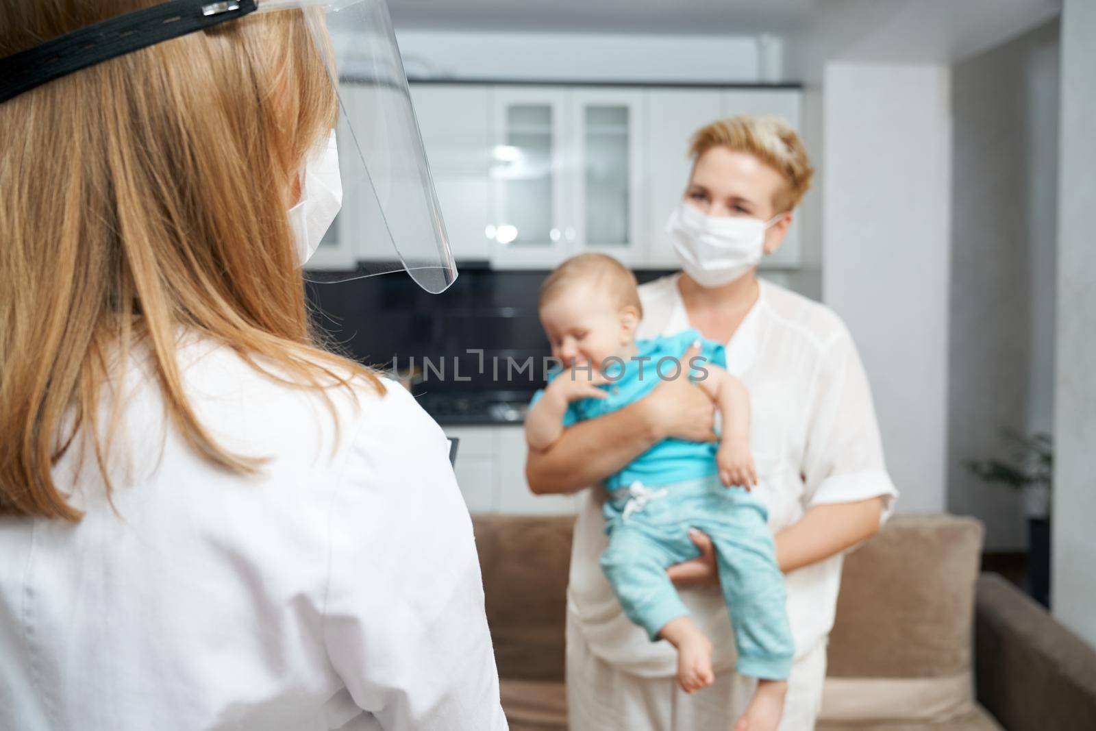 View from shoulder in medical uniform and mask talking with young woman that holding cute boy on hands. Health care at home.