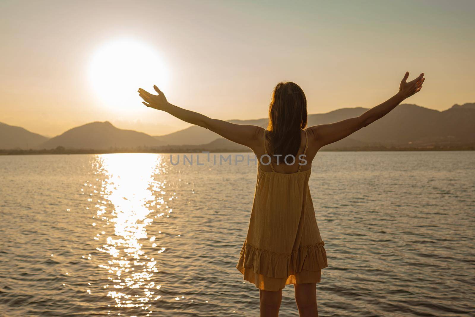 Lonely pensive woman with open arms in adoration of sun reflecting in ocean sea water at sunset or sunrise. Concept of spirituality and serenity in contact with nature to find yourself and one's soul