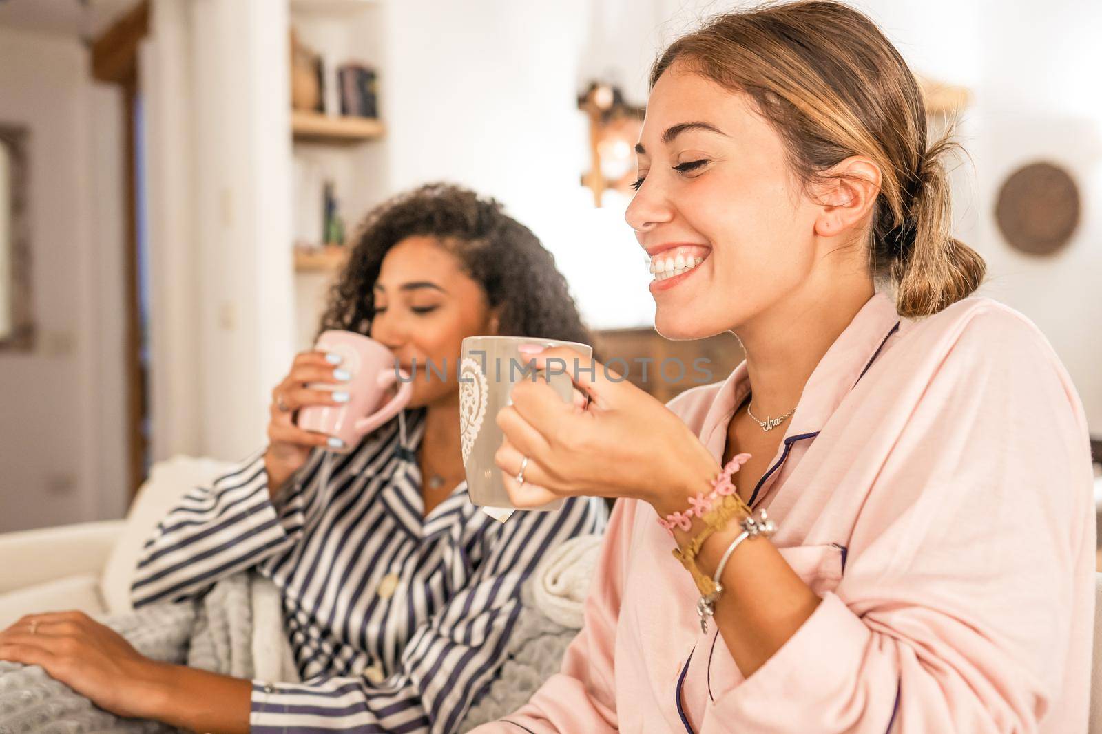 Couple of cohabiting girls in pajamas relaxing under blanket on cozy sofa drink a cup of tea while having fun talking and joking with each other. Real life moments of diverse multiracial gay couple by robbyfontanesi