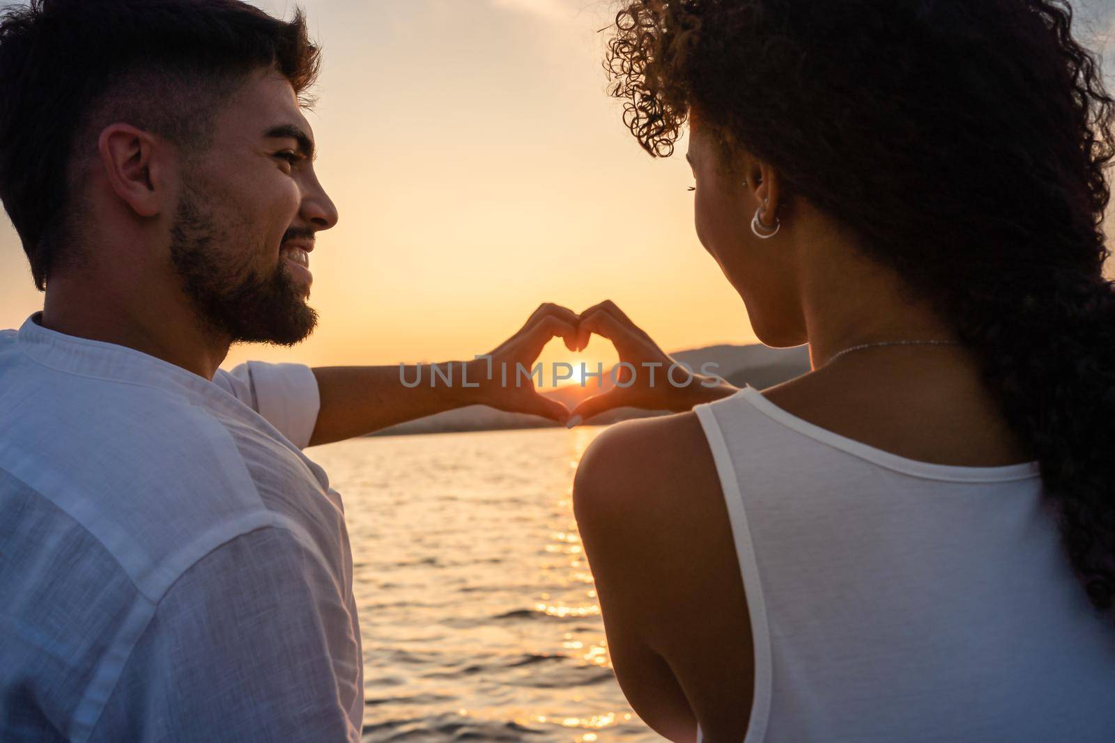 Young just married multiracial couple in love making heart shape with hands with setting sun in centre of it, looking in the eyes smiling. Romantic scene of Hispanic girl and boyfriend view from back by robbyfontanesi