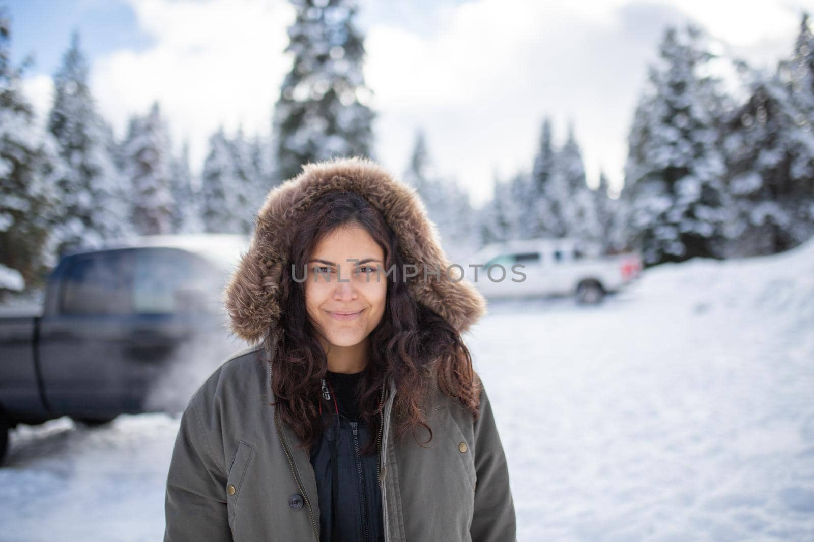 Happy woman standing with a beautiful snowy forest and trucks as background. Smiling hooded woman surrounded by snowy pine trees. Adventurous winter holiday