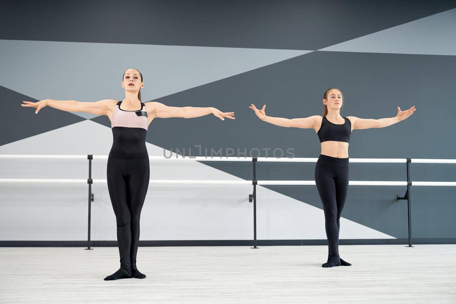 Adult female instructor helping young girl in black sportswear learn choreographic move. Two synchronized women practicing in hall, hi tech interior. Choreography, gymnastics concept.