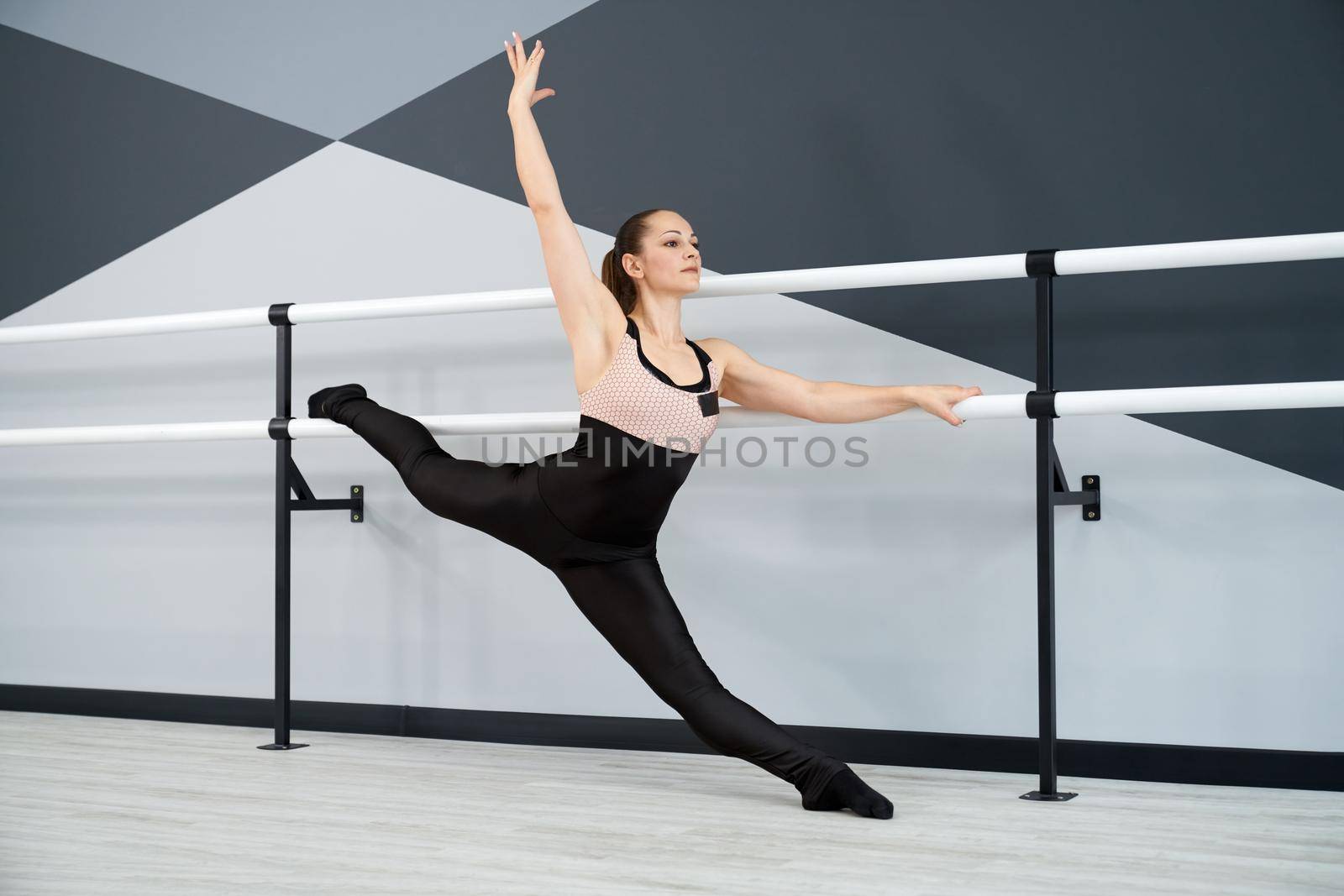 Side view of gorgeous brunette woman stretching leg on handrail, doing split. Fit flexible female athlete wearing sports outfit practicing in dance studio, hi tech interior. Concept of gymnastics.