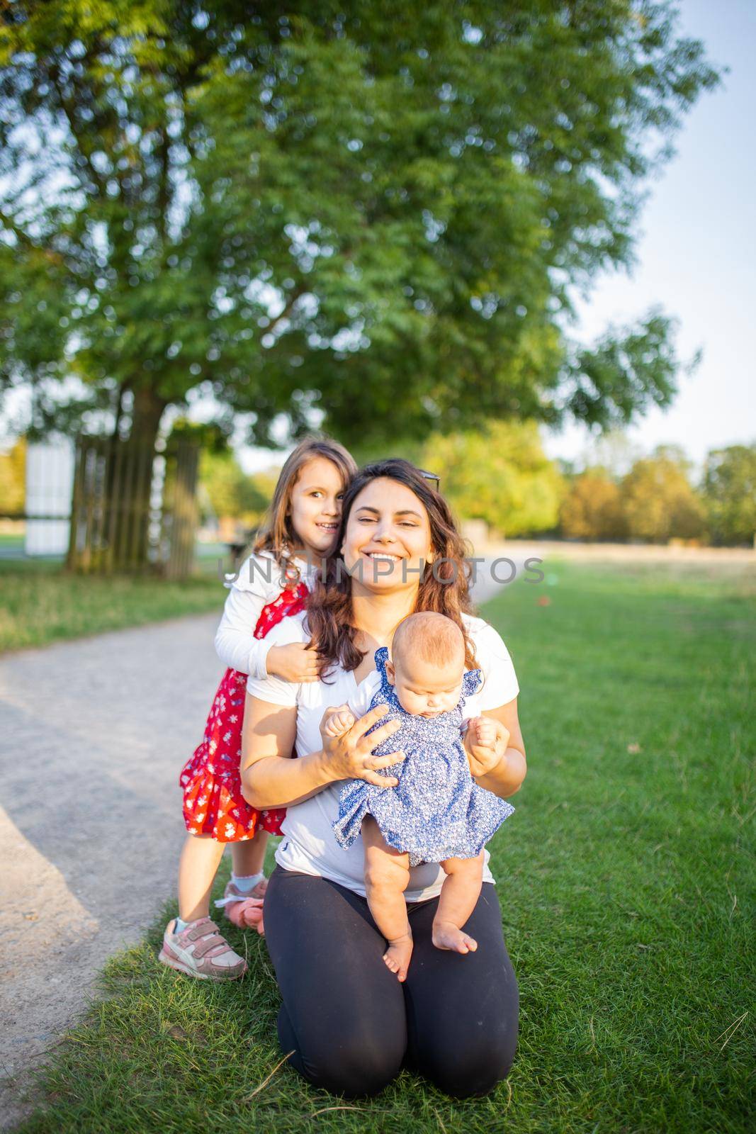 Happy woman, baby, and little girl sitting on grass next to a path and with trees as background. Smiling mother playing with adorable daughters in park. Young family having fun outdoors