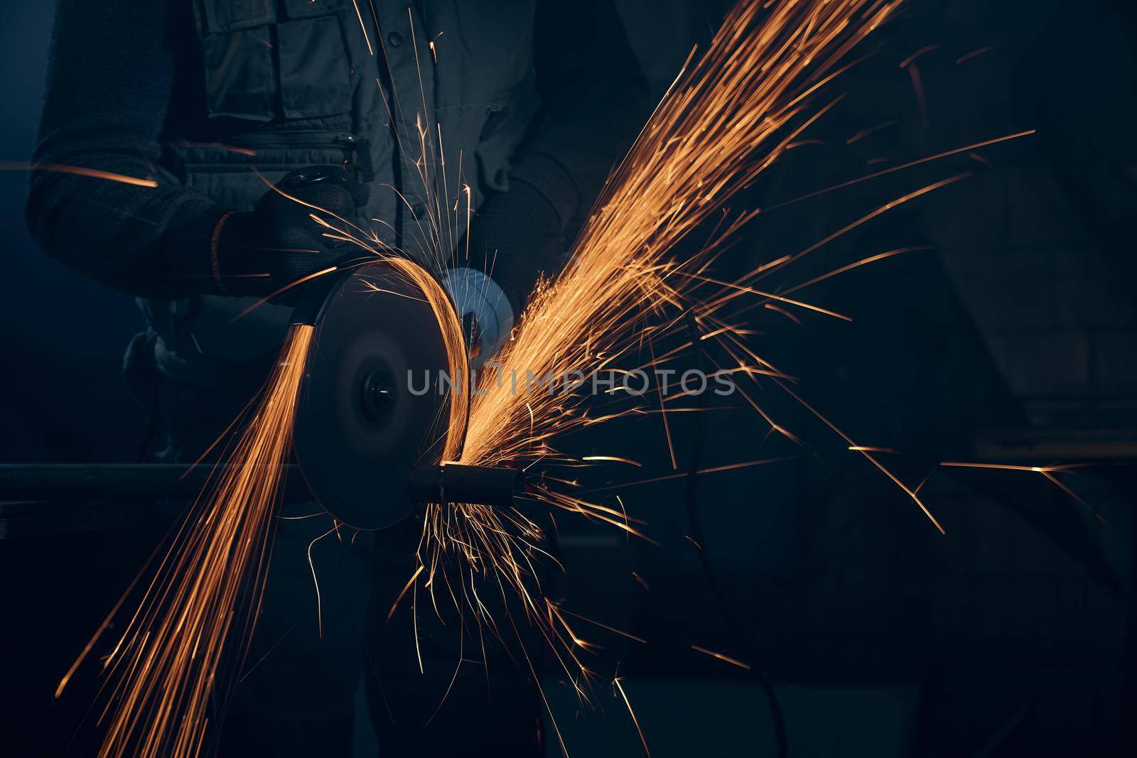 Close up of worker in special black suit working with angle grinder and grinding metal with large yellow sparks. Concept of polishing metal with special equipment in dark room.