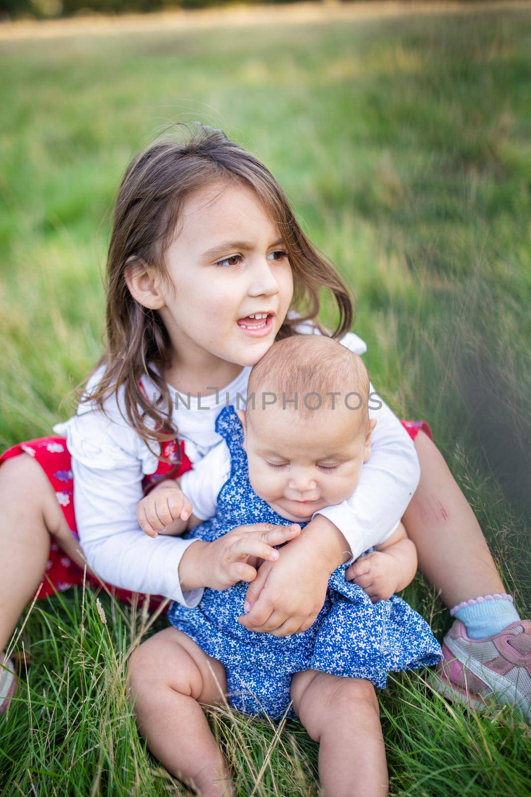 Adorable little girl sitting on grass and lovingly hugging baby. Portrait of cute young child sitting with her baby sibling in park. Young family playing outdoors