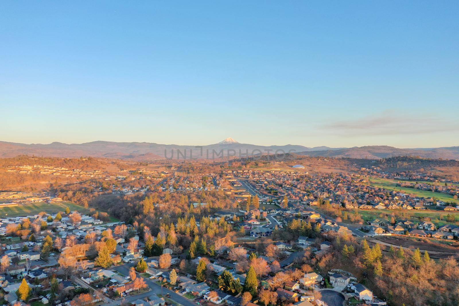 Peaceful town surrounded by trees with mountains as background during beautiful sunset. Aerial view of houses surrounded by autumn-colored nature. Rural town landscape