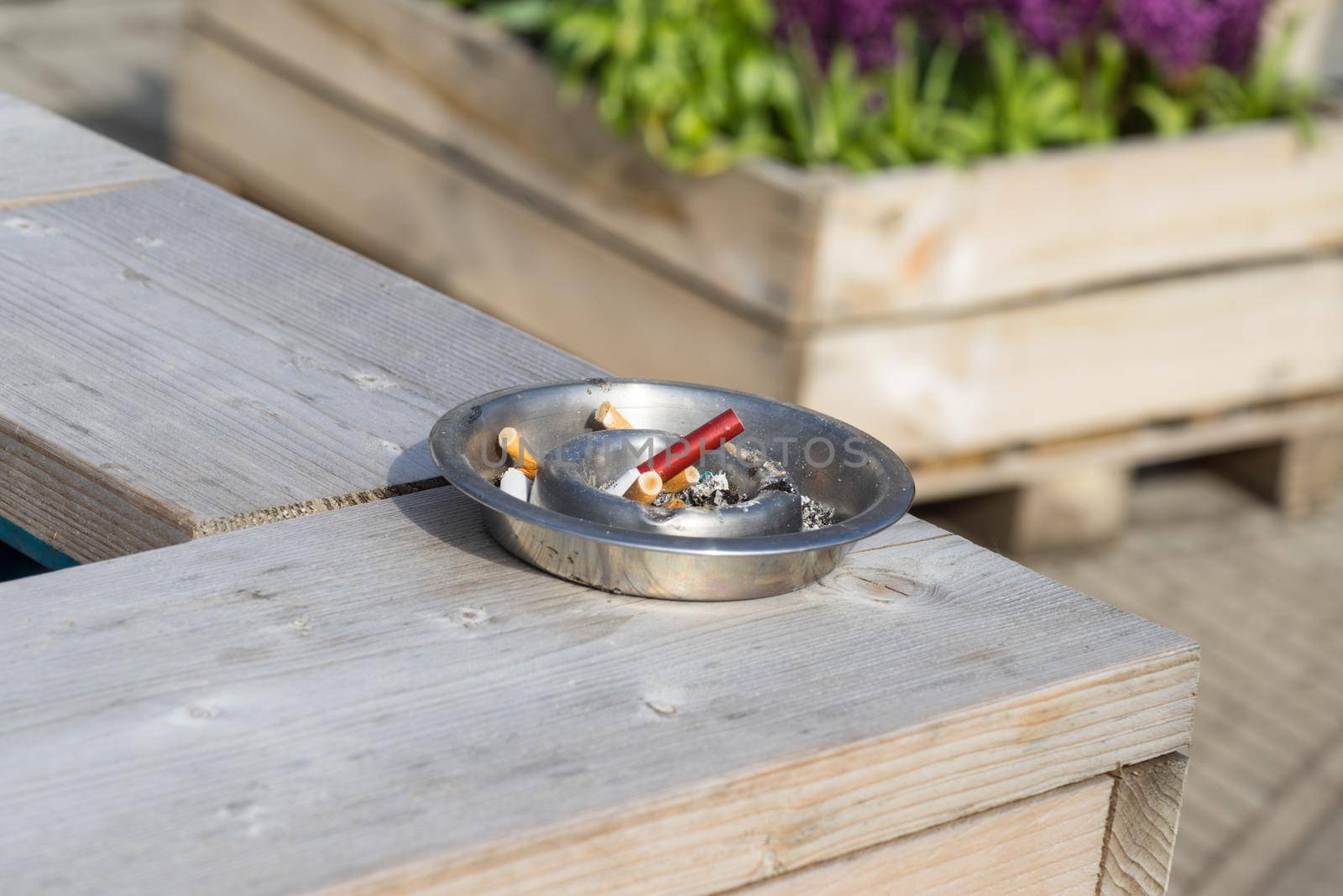 silver ashtray with cigarette butts on a wooden table outside on an sunny summer day by LeoniekvanderVliet