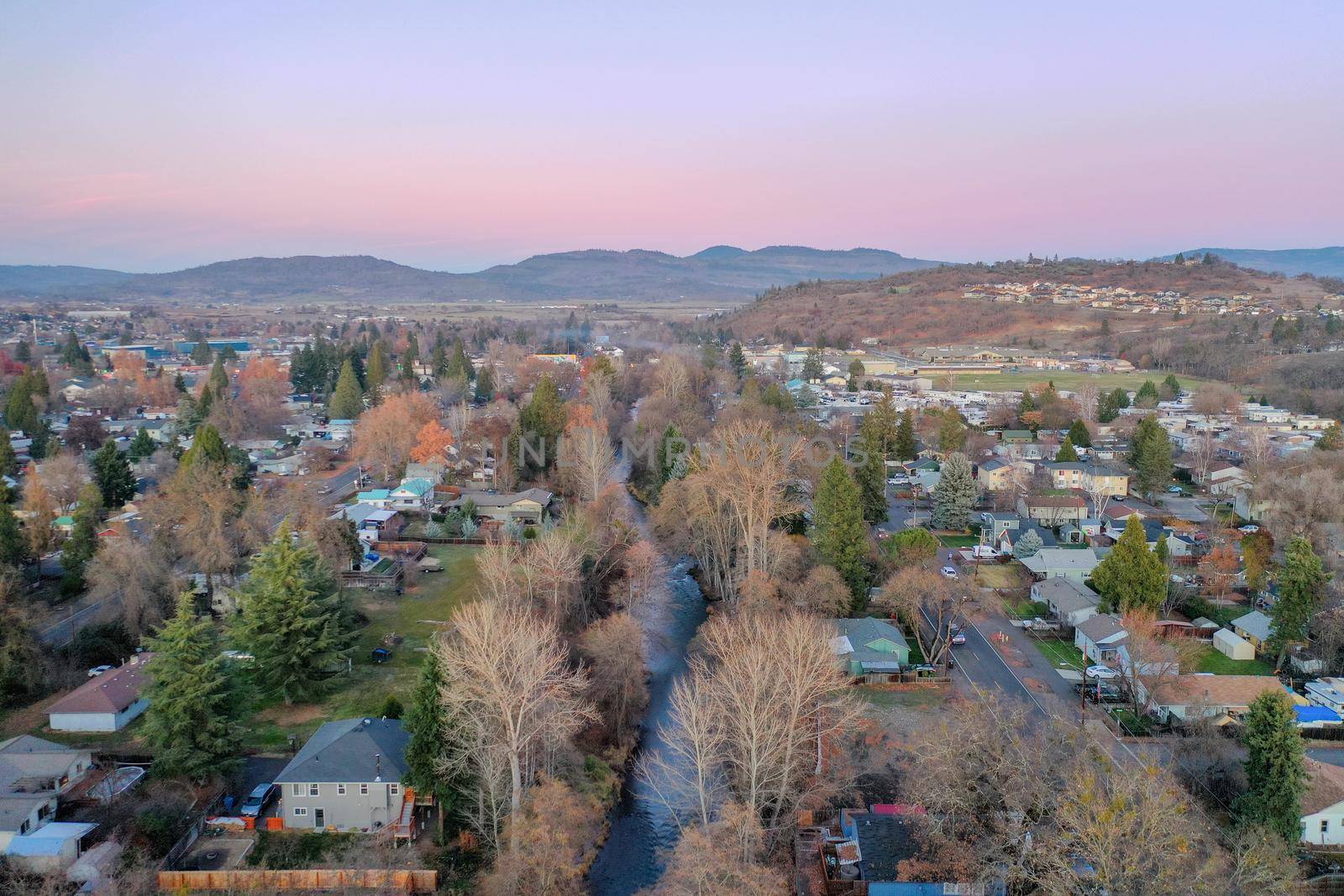 Peaceful town surrounded by trees with mountains as background during sunset. Aerial view of roads and houses surrounded by autumn-colored nature. Rural town landscape