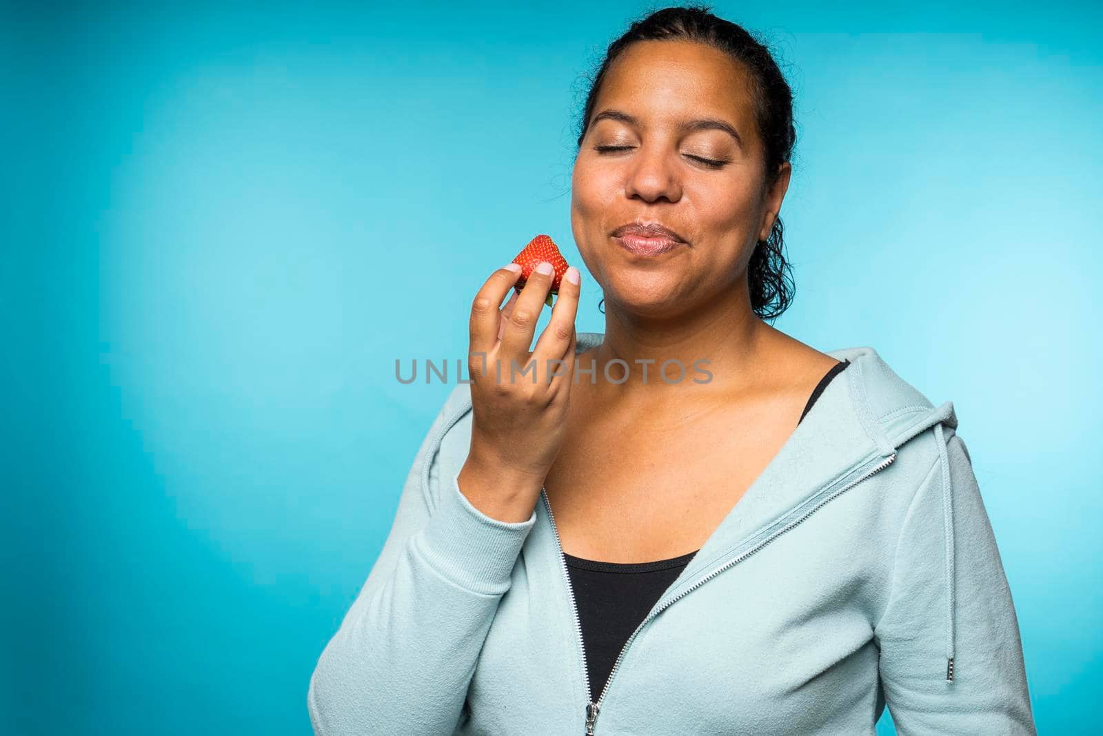 Beautiful young mixed race woman in casual clothing eating and enjoying a fresh strawberry with a blue background by LeoniekvanderVliet