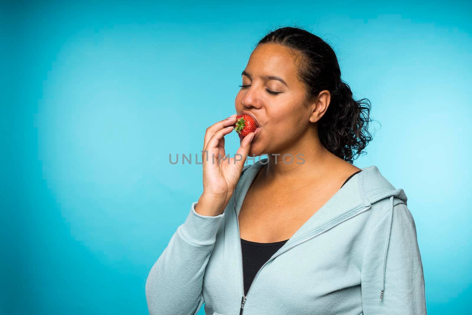 Beautiful young mixed race woman in casual clothing eating and enjoying a fresh strawberry with a blue background by LeoniekvanderVliet