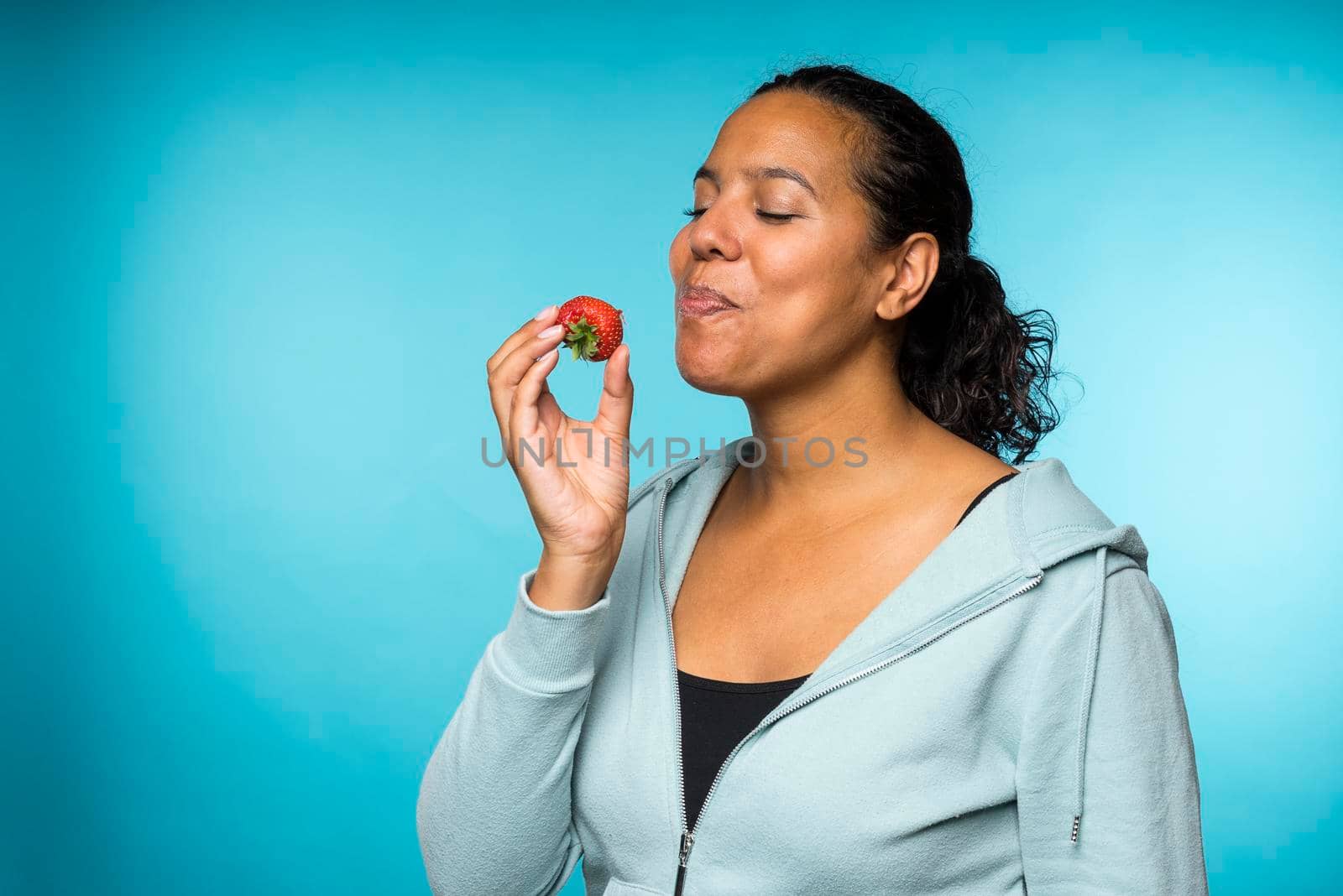Beautiful young mixed race woman in casual clothing eating and enjoying a fresh strawberry with a blue background by LeoniekvanderVliet