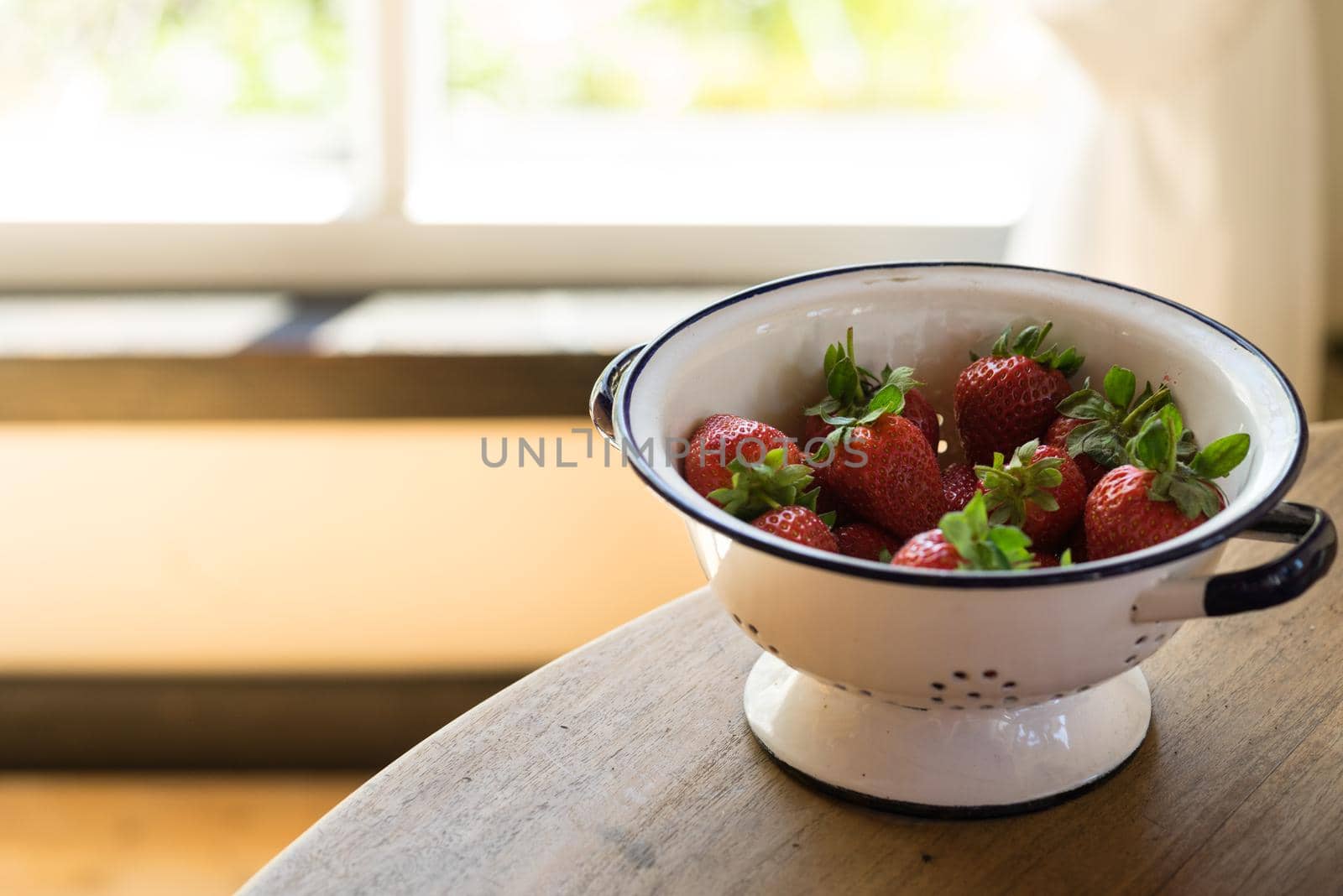 Bowl of fresh strawberries in a white bowl or colander on a table in front of a window