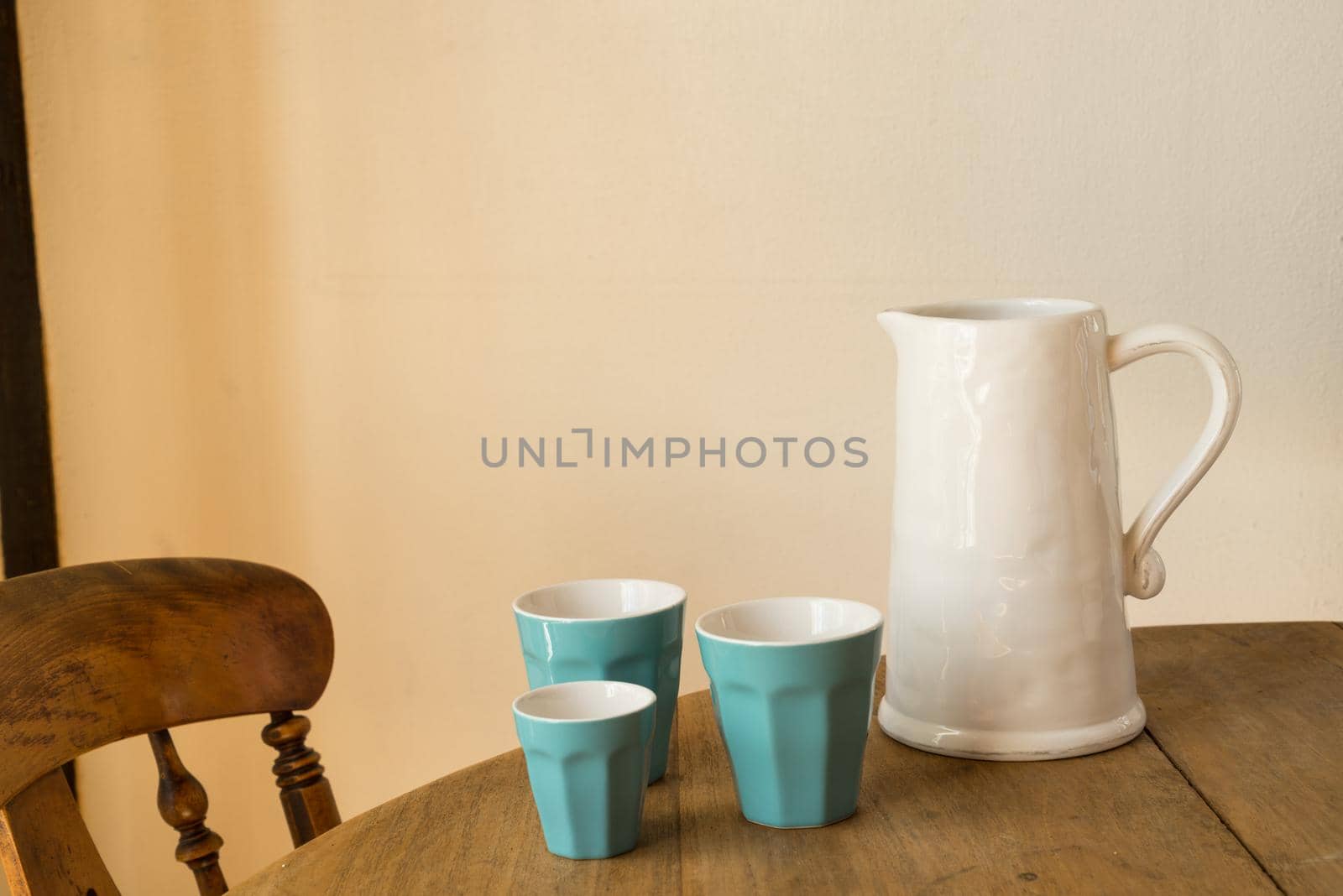 white jar or jug with blue cups on a wooden table with a chair by LeoniekvanderVliet