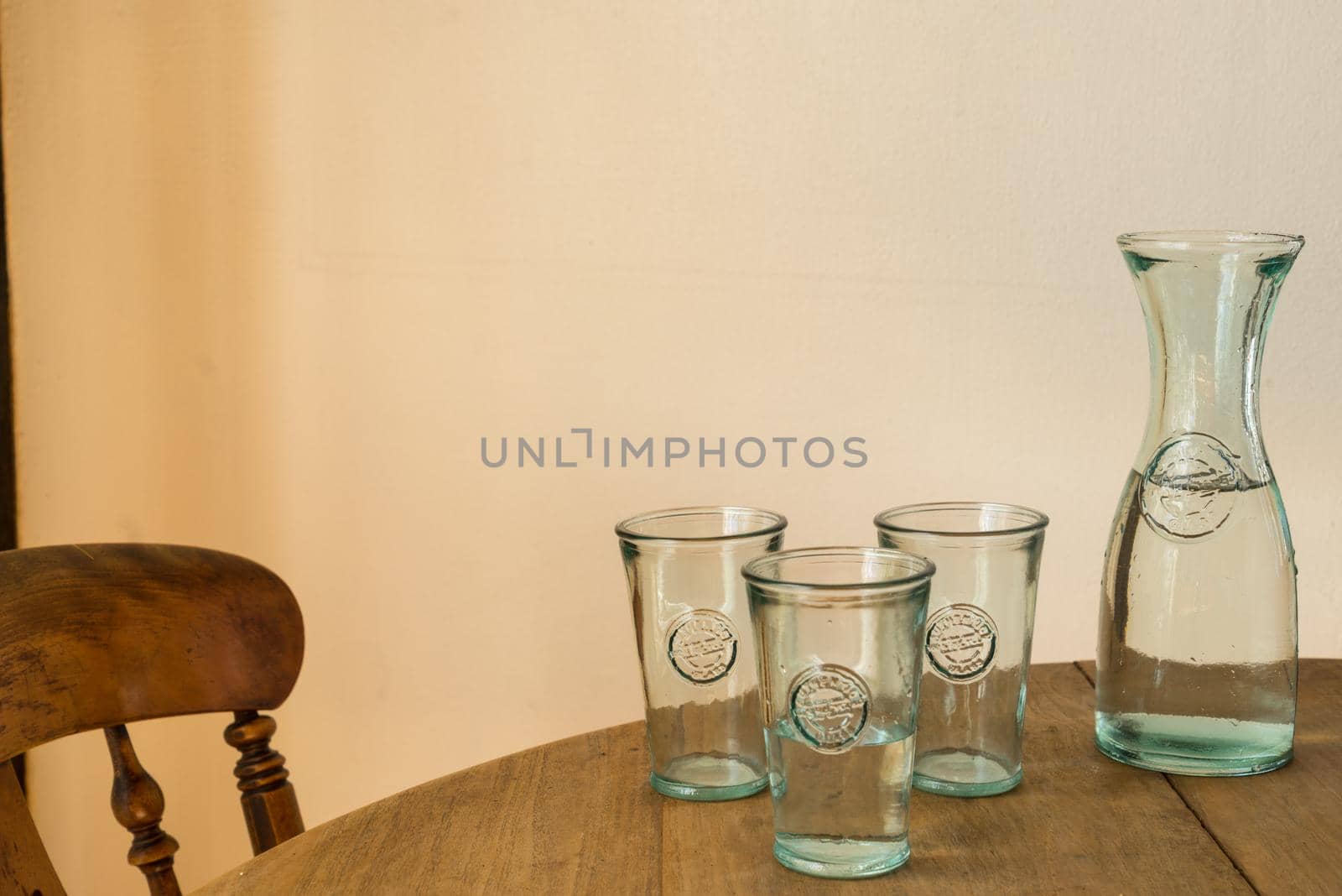 glass bottle or jar with water with three glasses in a wooden table with a chair indoors by LeoniekvanderVliet