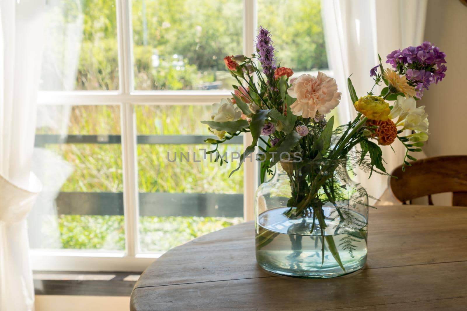 Vase with a mixed bouquet of flowers standing on a wooden table behind a window in the kitchen by LeoniekvanderVliet