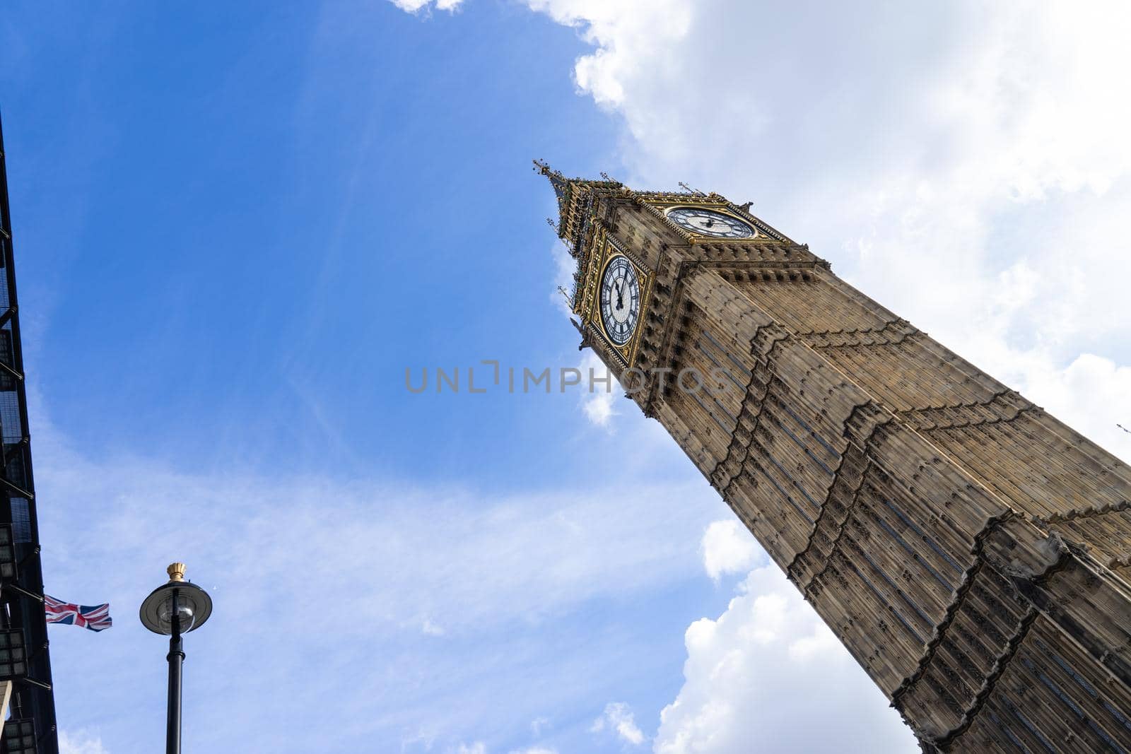 Low angle view of the Big Ben in London England United Kingdom against a blue sky with white clouds and a Union Jack British flag on a building and a streetlight by LeoniekvanderVliet