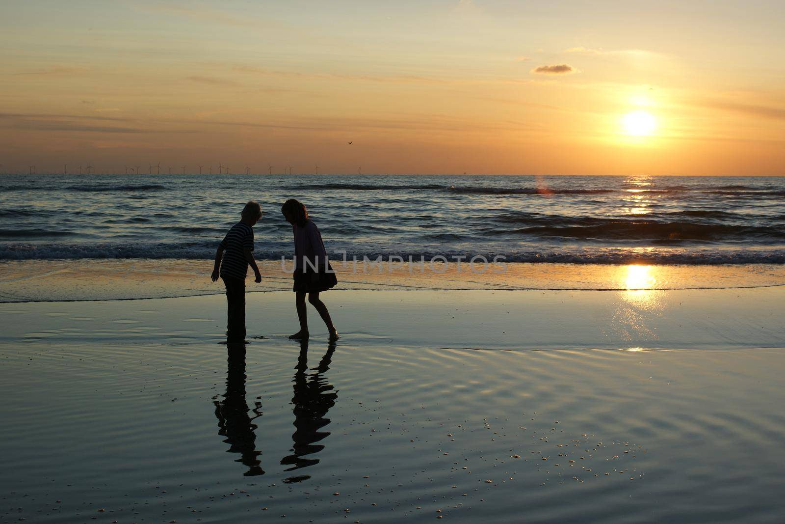 Silhouettes of two anonimous children against a sunset sky at the beach with reflection and windmills at the horizon by LeoniekvanderVliet