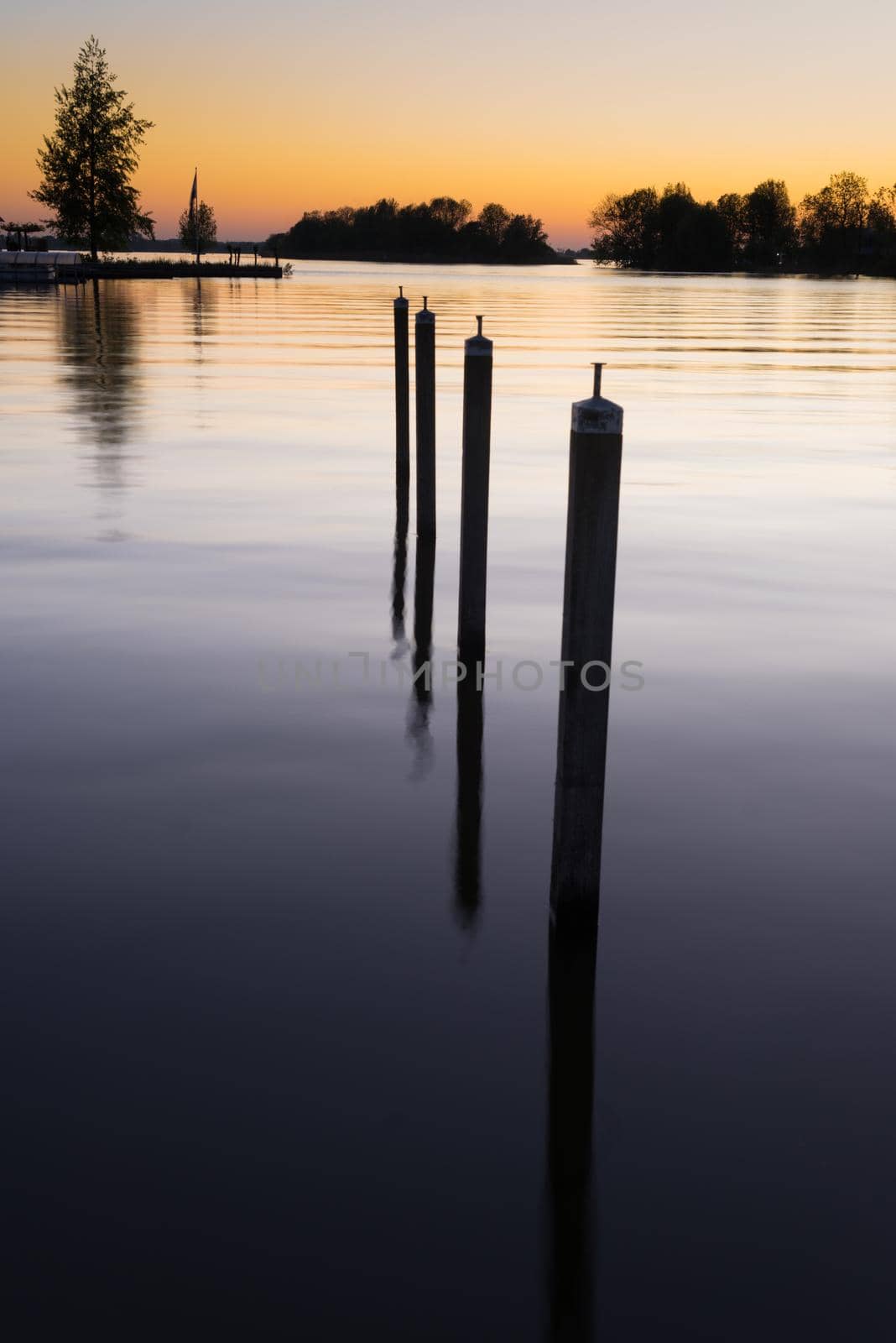 Row of poles in smooth water on the shore of a lake at dusk showing the reflection of sunset in the water a concept of perspective or the fall of the evening by LeoniekvanderVliet