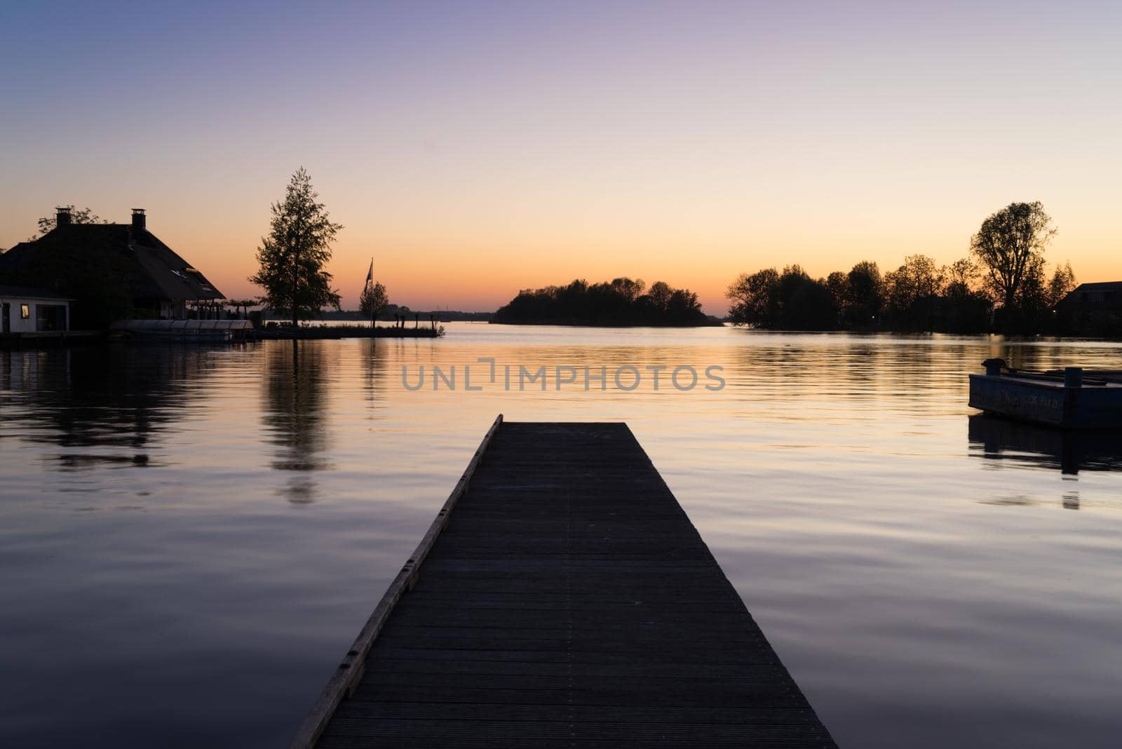 Pier in smooth water on the shore of a lake at dusk showing the reflection of sunset in the water a concept of perspective or the fall of the evening by LeoniekvanderVliet