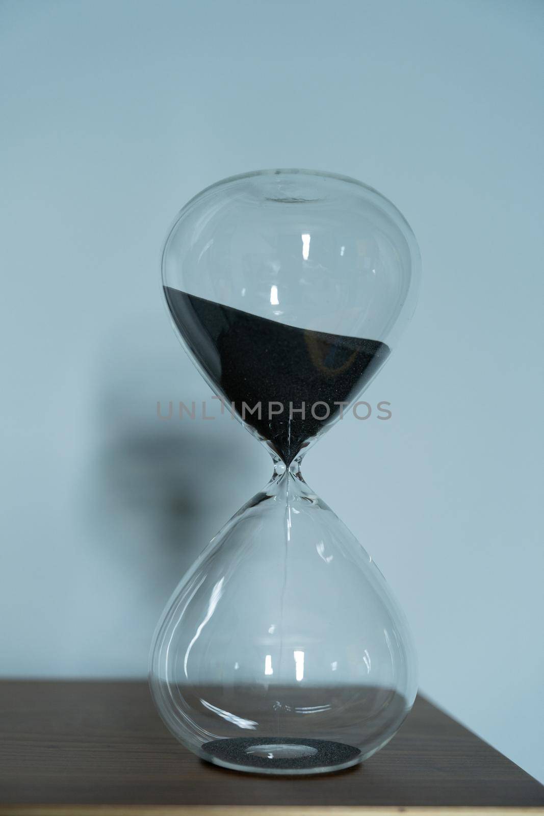 Black sand running through the bulbs of hourglass measuring the passing time in a countdown to a business deadline, urgency and running out of time on a blue background with copy space.