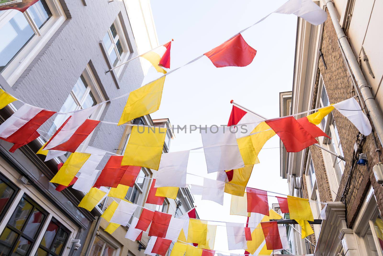 Dutch square flags on a ribbon, in red, white and yellow, of traditional festival named Carnaval, like Mardi Gras, in 's-Hertogenbosch, Oeteldonk with a blue sky  by LeoniekvanderVliet