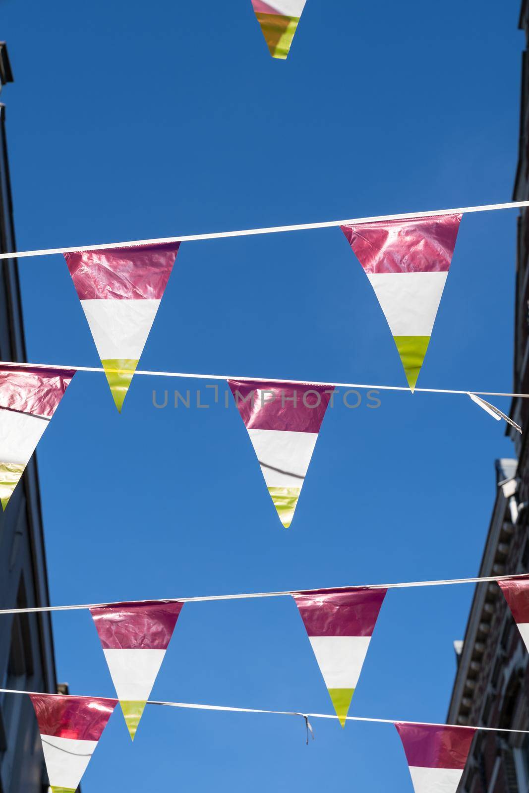 Dutch triangular flags on a ribbon, in red, white and yellow, of traditional festival named Carnaval, like Mardi Gras, in 's-Hertogenbosch, Oeteldonk with a blue sky  by LeoniekvanderVliet